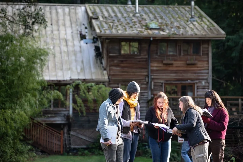 Six people standing outdoors in front of a wooden house, discussing and looking at documents or notebooks.