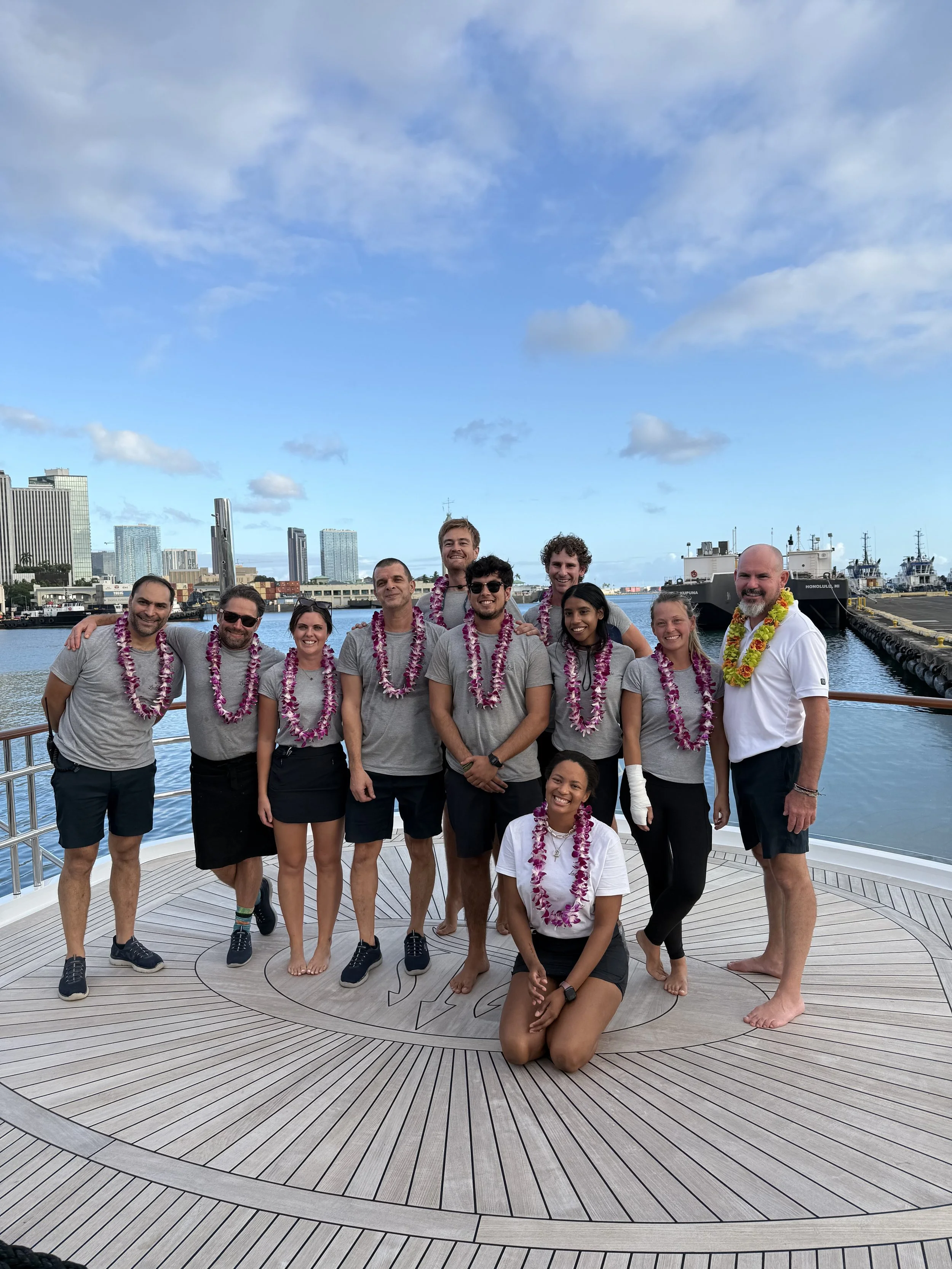 Group of eleven people wearing leis and casual clothing on a boat deck with city skyline and boats in the background.