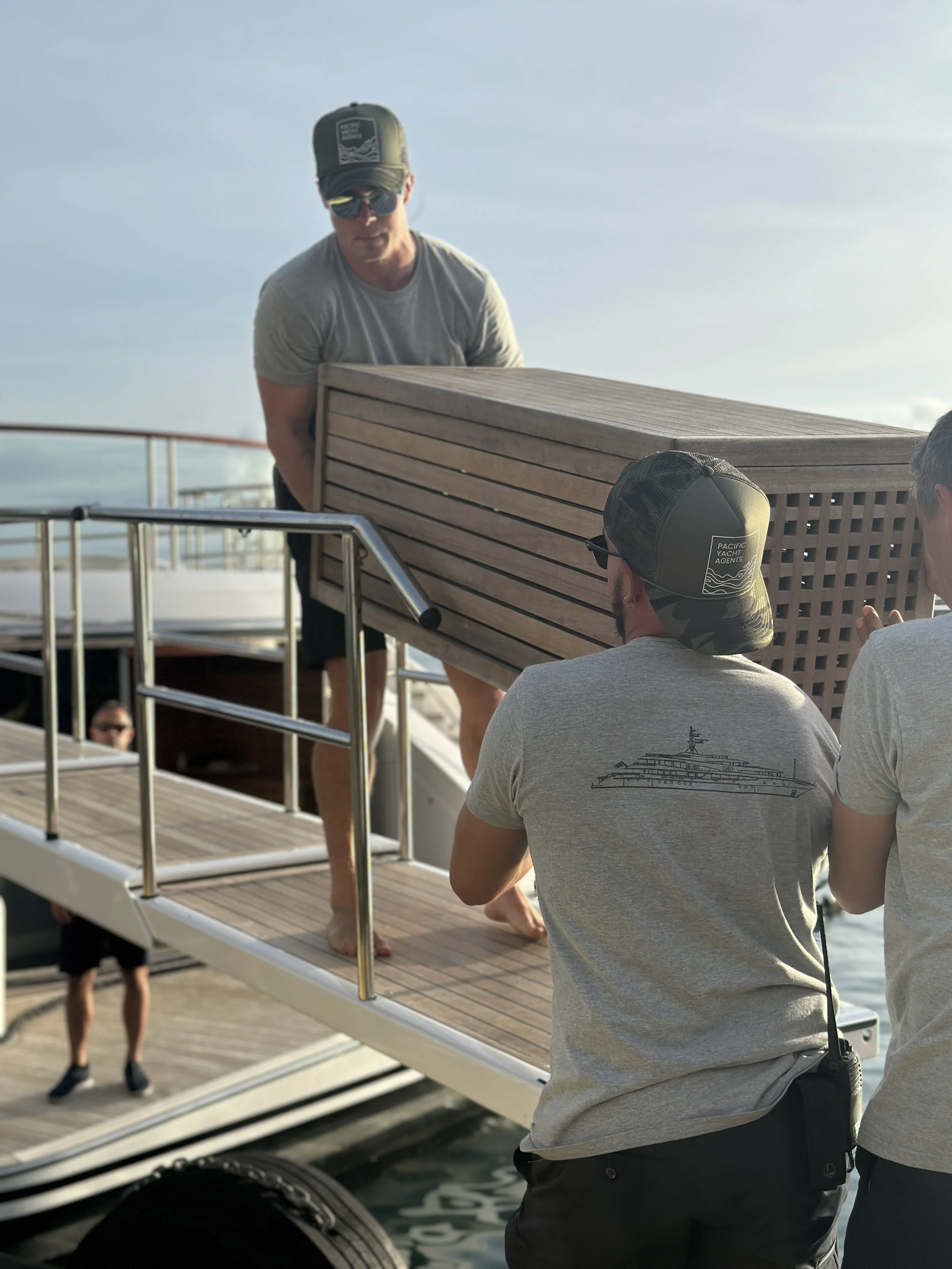 People unloading a wooden hot tub from a boat onto a dock by the water, with some individuals wearing sunglasses and caps.