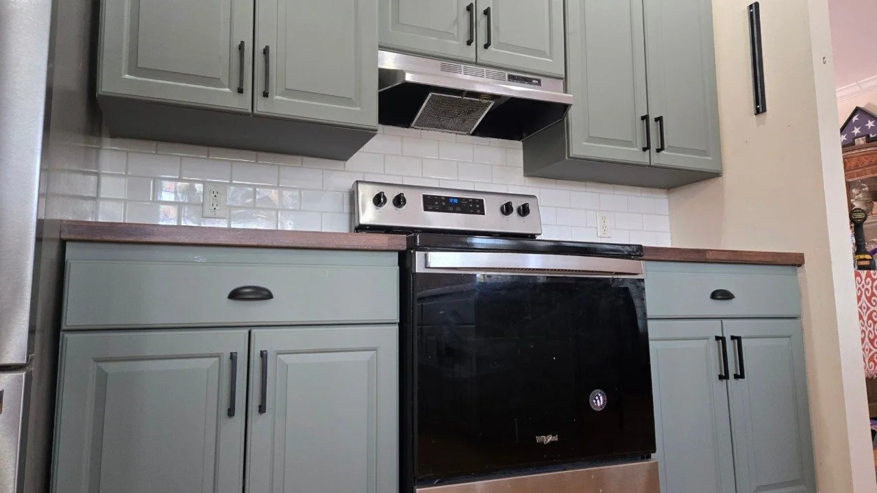 Photo of a kitchen with gray cabinets, a black stove, white subway tile backsplash, and a wooden countertop.