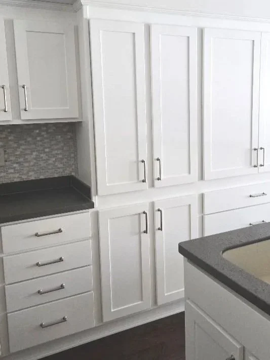 White kitchen cabinets with silver handles, gray countertops, a small section of backsplash, and a gray sink in the foreground.