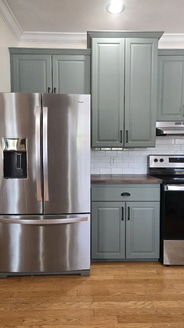 Kitchen with light blue cabinets, stainless steel refrigerator, white tile subway backsplash, and black stove on wooden floor.