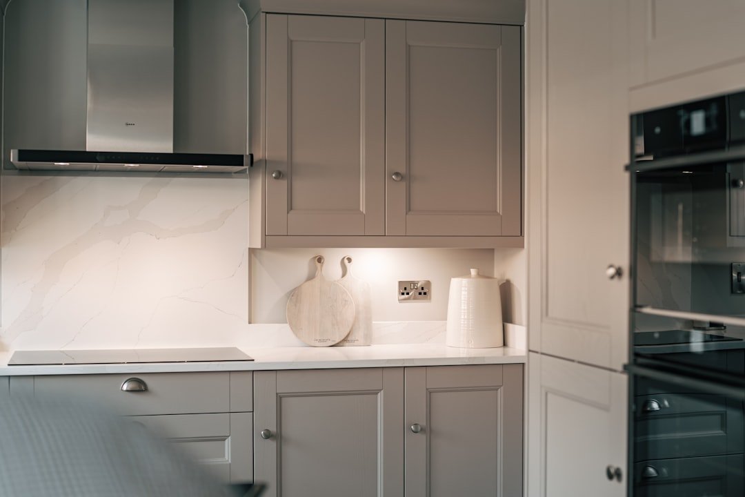 Kitchen with gray cabinets, marble backsplash, and white countertop. Two wooden cutting boards with handles and a ceramic container are on the counter, under a spotlight. There is an electric outlet and a stainless steel range hood.
