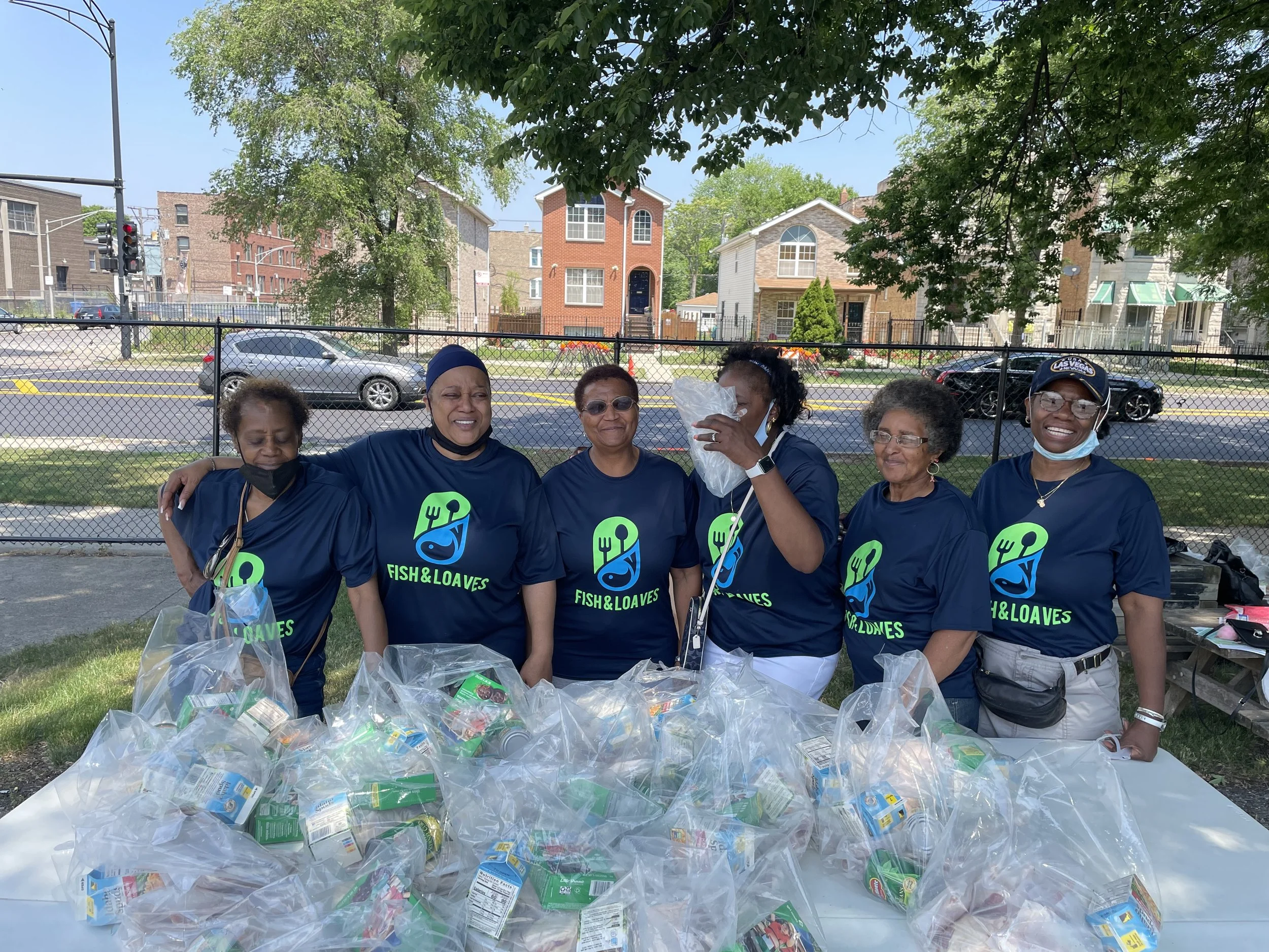 Group of people wearing "Fish & Loaves" T-shirts standing outside behind a table with food packages, trees and buildings in the background.