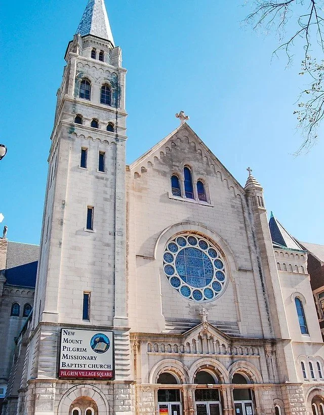 Front view of New Mount Pilgrim Missionary Baptist Church with a tall steeple and round stained glass window.