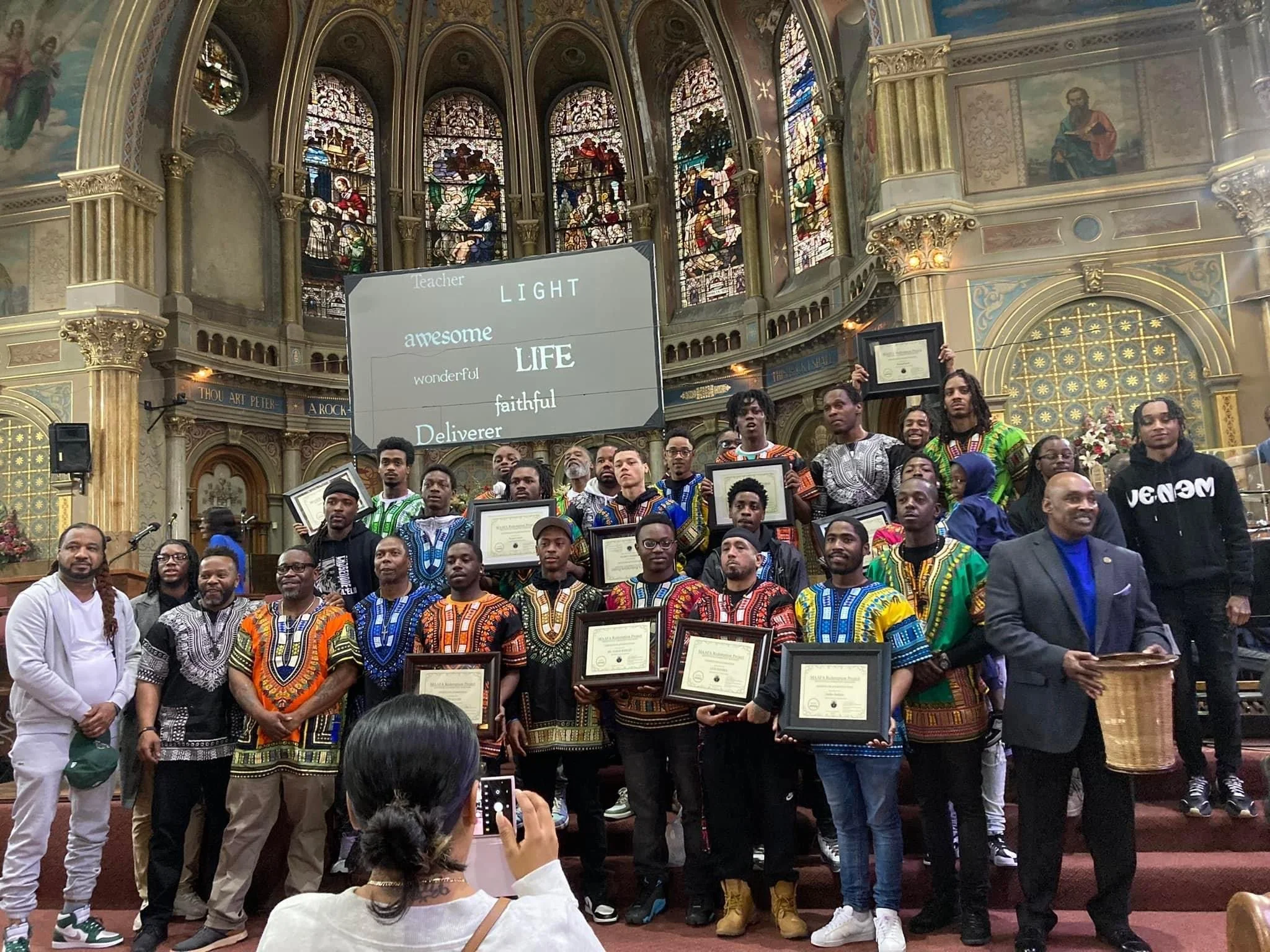 A group of people stands on a stage inside a church, holding framed certificates. Many are wearing colorful traditional garments. Stained glass windows are visible in the background, along with a large screen displaying words like "awesome," "wonderful," and "faithful." The setting is ornate, with detailed architectural elements.