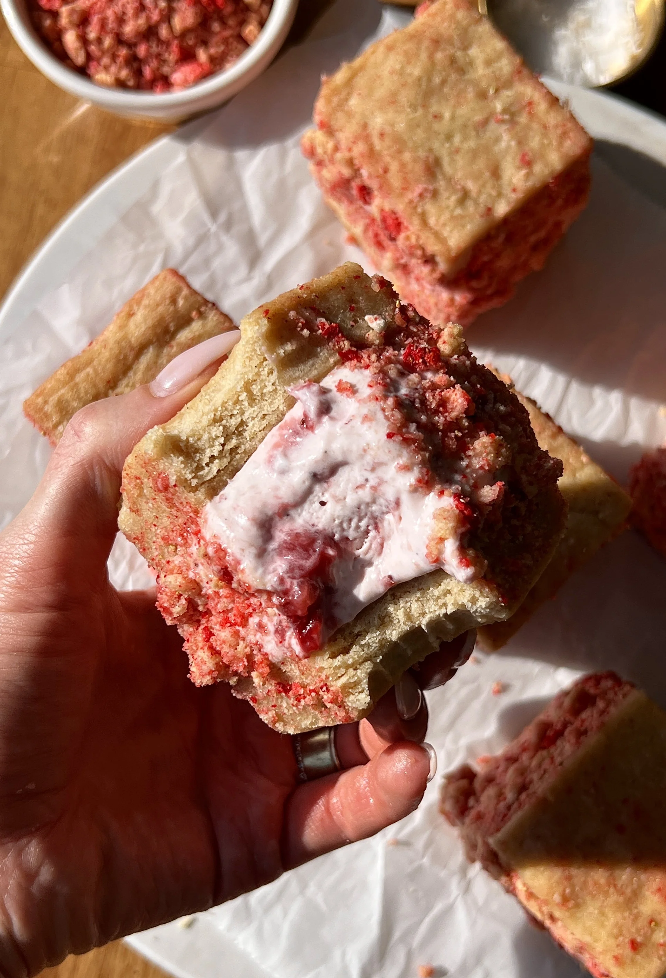 Strawberry shortcake shortbread ice cream sandwich with a bite taken out showing the soft buttery shortbread layers with rich and creamy homemade no-churn strawberry ice cream and strawberry shortcake crunch coating inside.