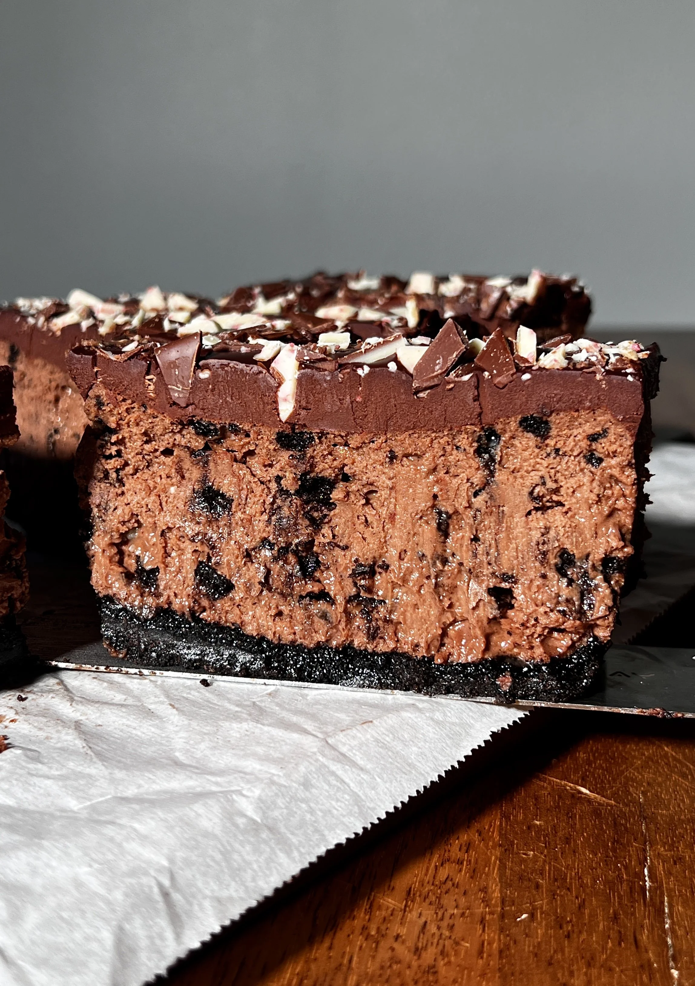 Close-up of peppermint Oreo cheesecake bars with a dark Oreo crust, creamy peppermint mocha cheesecake filling, chocolate ganache topping, and chopped peppermint bark sprinkled on top.