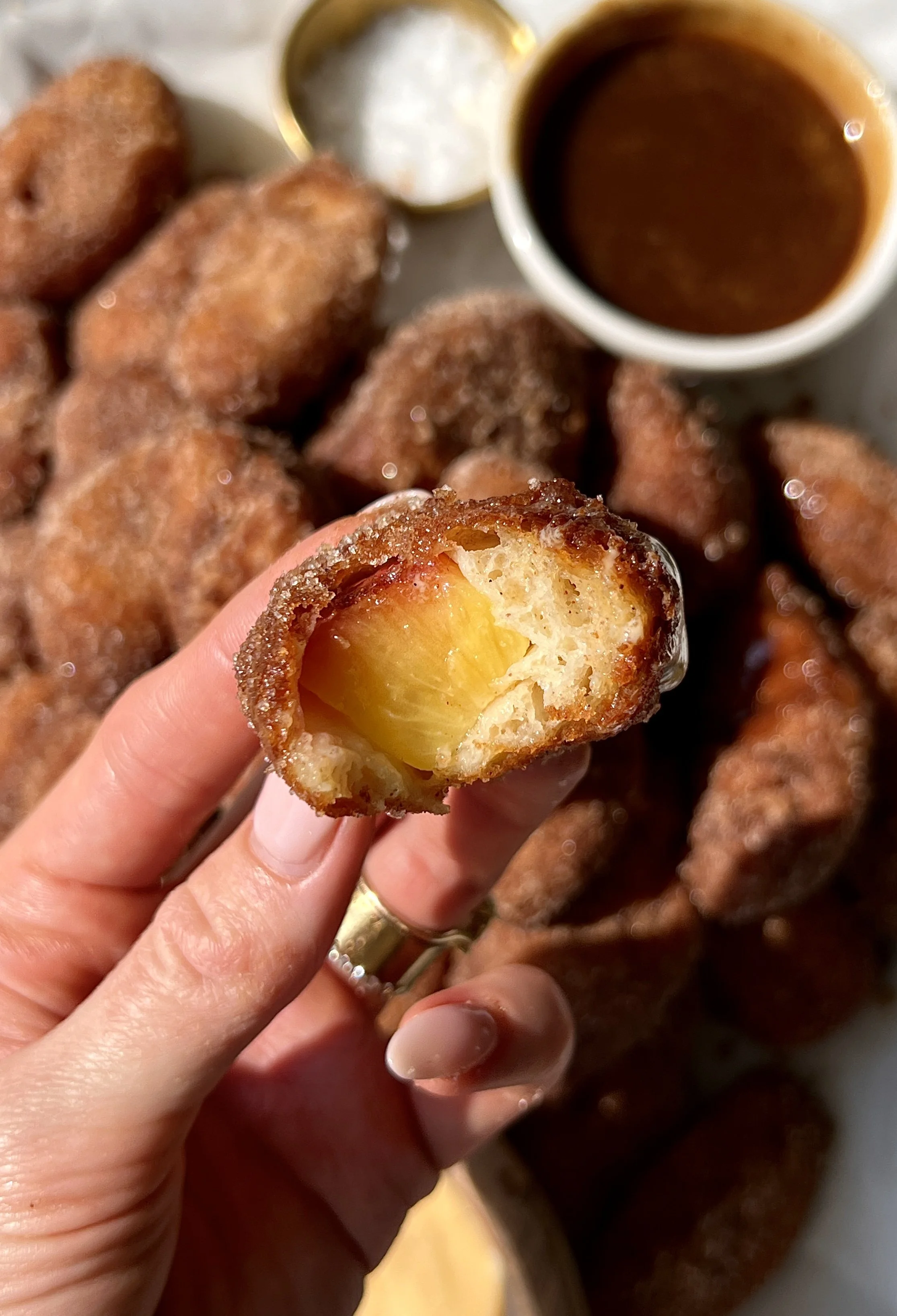 Buttermilk fried peach rolled in cinnamon sugar with a bite taken out showing the golden crisp exterior and warm juicy peach inside served with homemade crème anglaise and salted caramel.