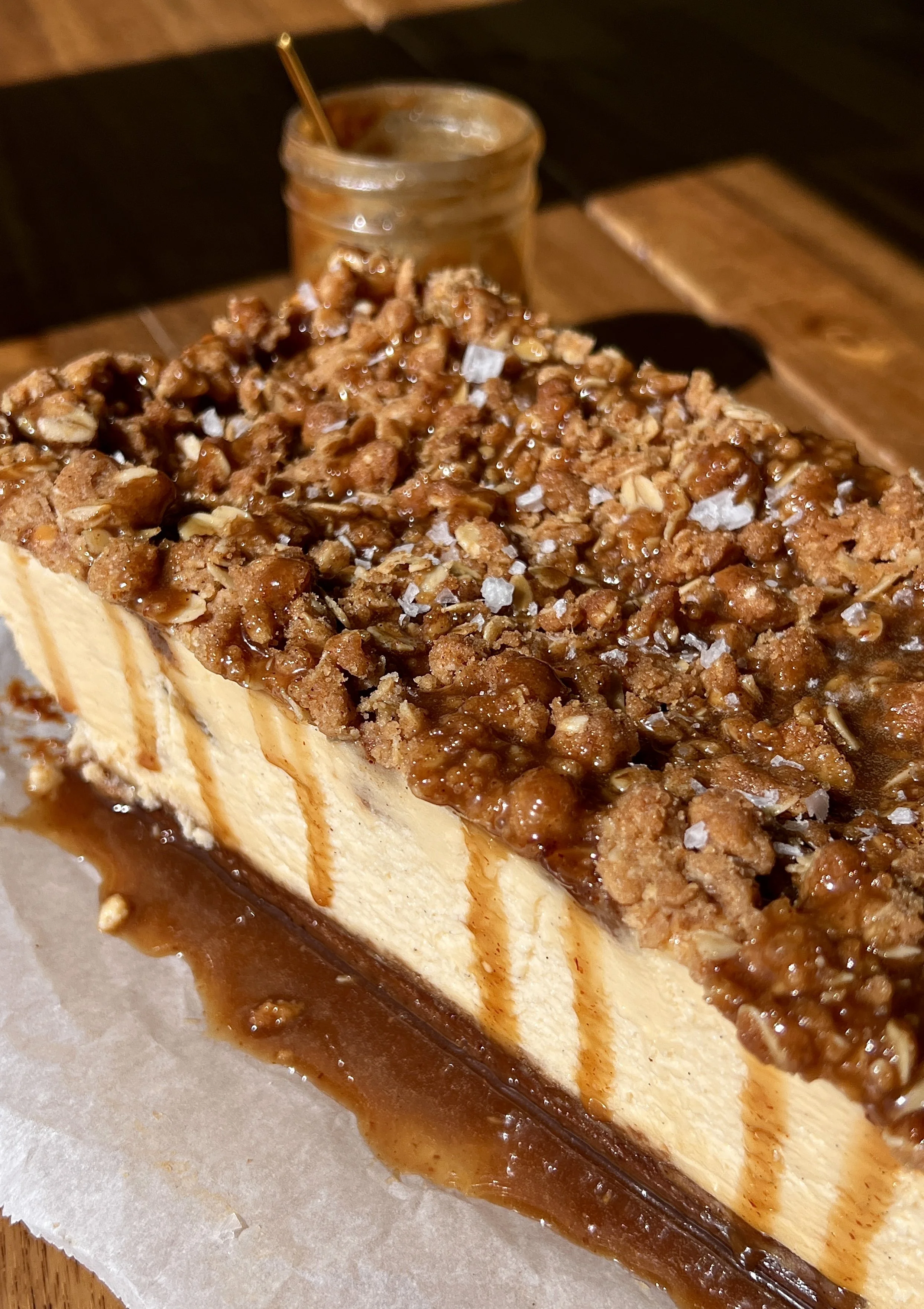 Close-up of a salted caramel apple crumble cheesecake bar showing the golden Biscoff cookie crust, rich and creamy vanilla bean cheesecake filling, warm gooey spiced apple pie filling, and golden crumble topping throughout.