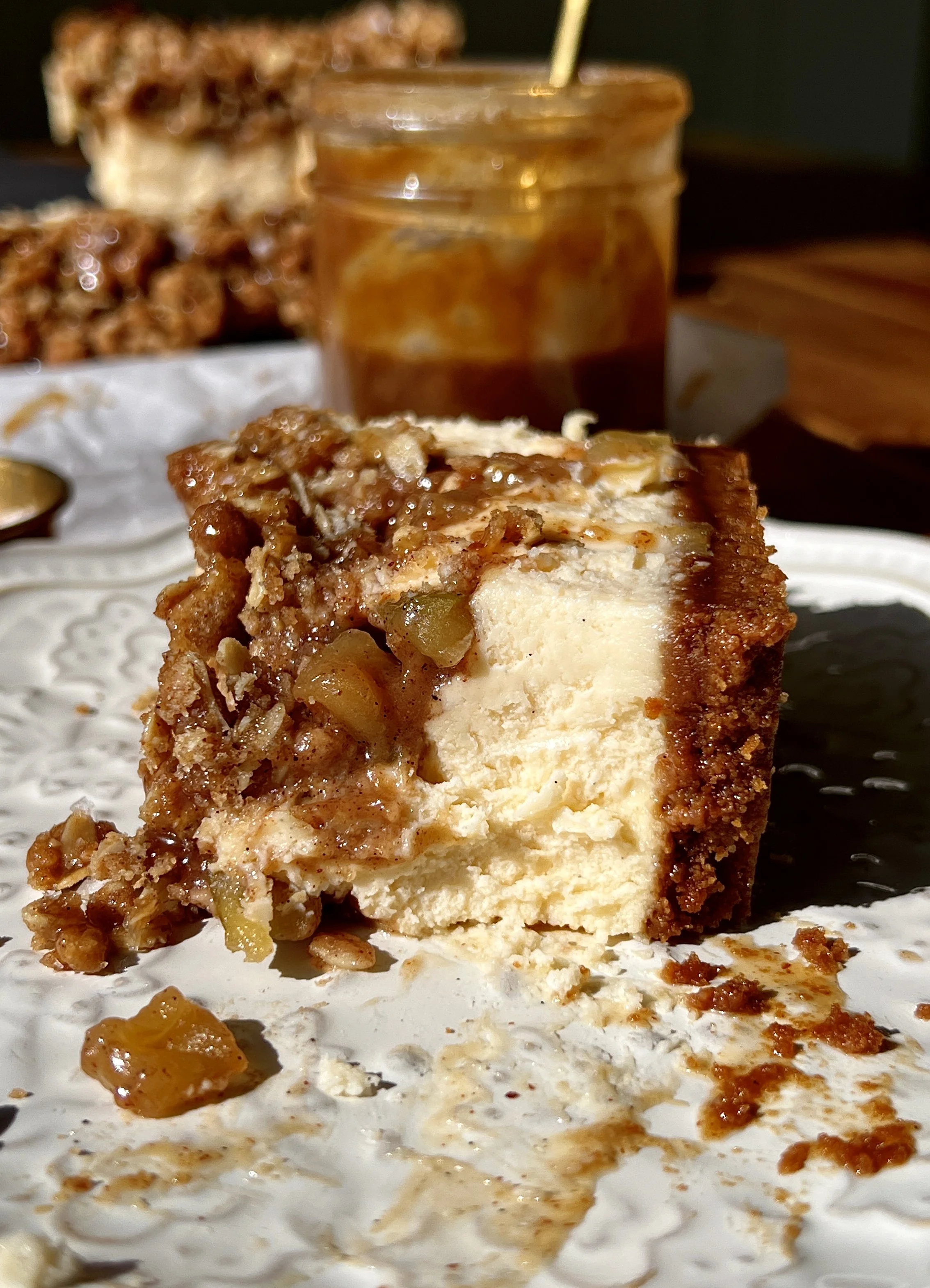 Close-up of a salted caramel apple crumble cheesecake bar showing the golden Biscoff cookie crust, rich and creamy vanilla bean cheesecake filling, warm gooey spiced apple pie filling, and golden crumble topping throughout.