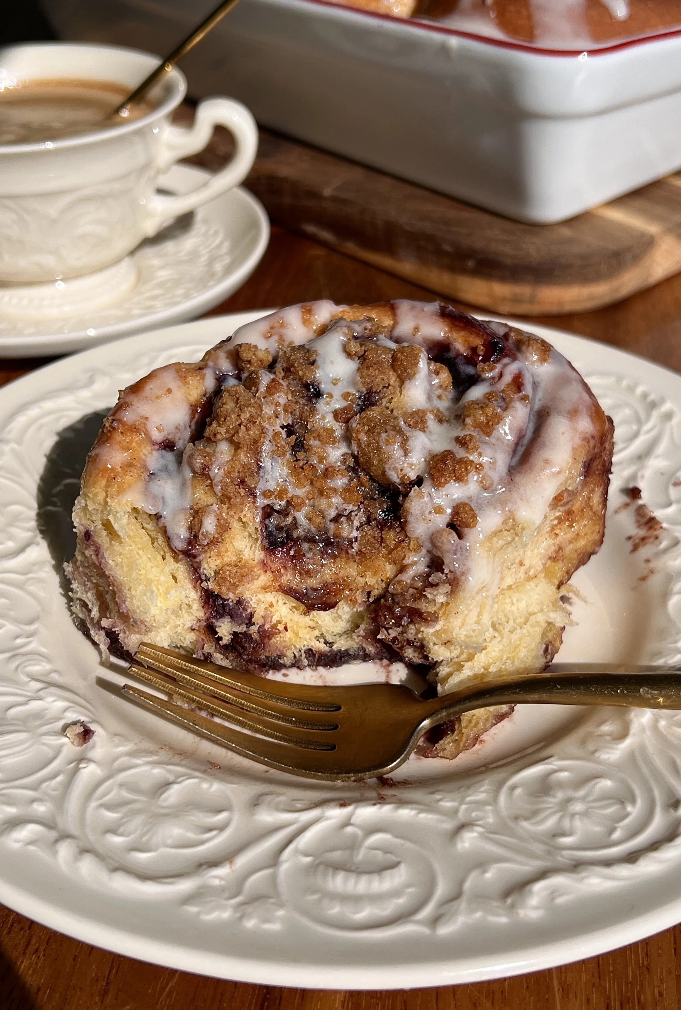 Warm blueberry crumble cinnamon rolls served with cream cheese glaze and a cup of coffee.