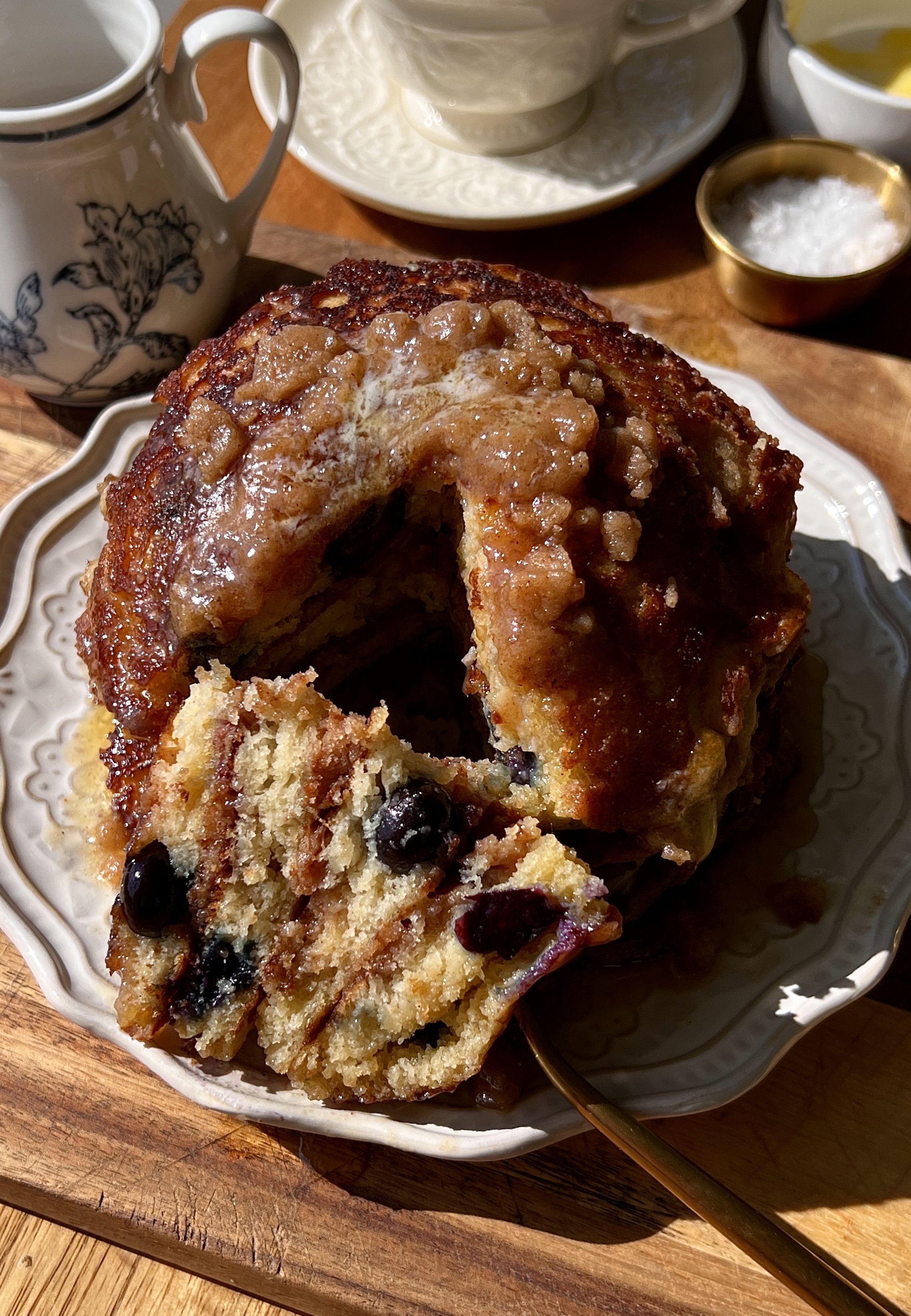 Stack of golden brown butter blueberry muffin pancakes with caramelized cinnamon streusel and juicy blueberries served warm with a pat of butter, maple syrup, and a hot cup of coffee.