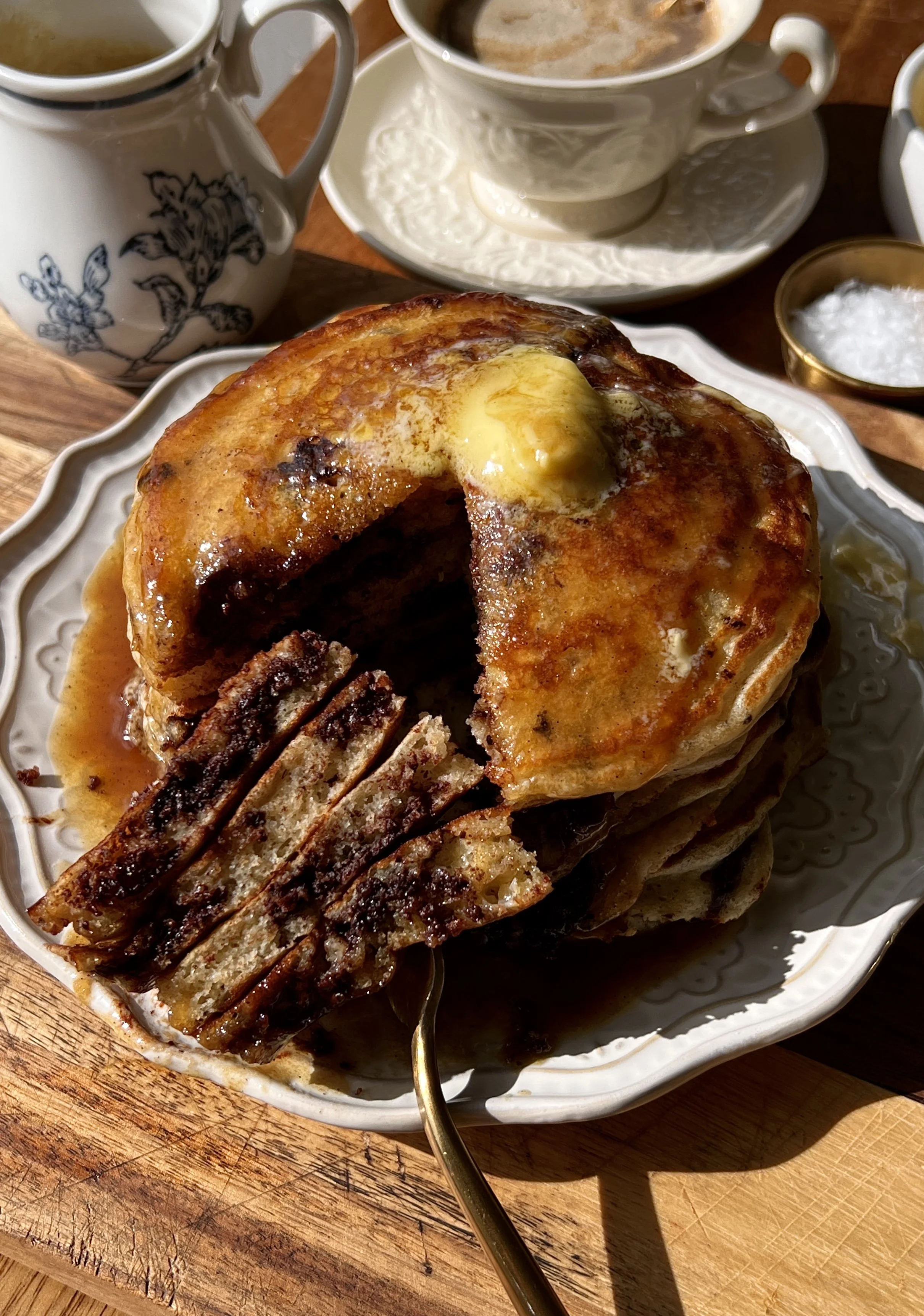 Stack of golden brown butter chocolate chip pancakes with maple espresso buttermilk syrup drizzled on top served with a hot cup of coffee.