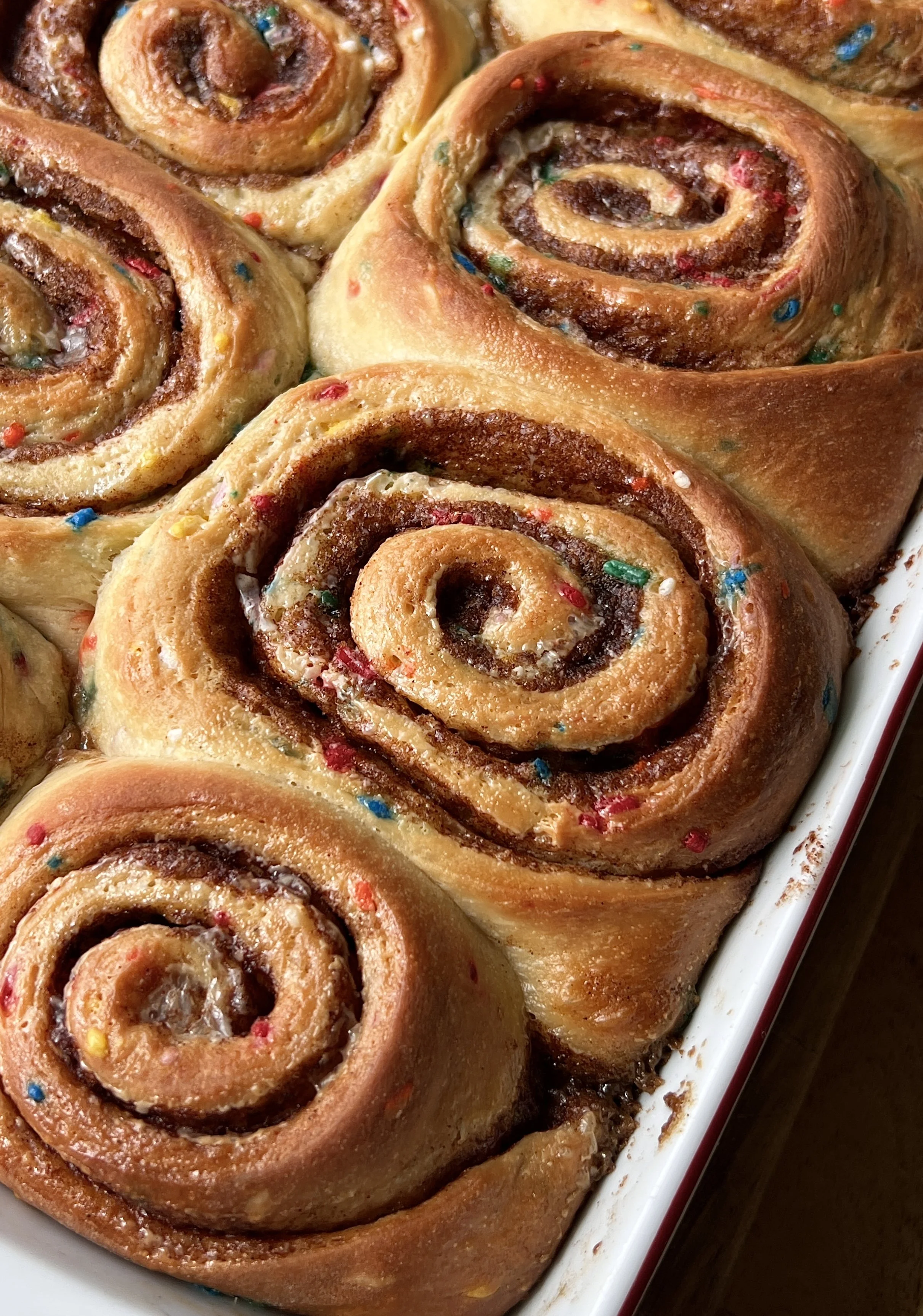 Pan of golden funfetti cinnamon rolls with sticky caramelized edges topped with smooth cream cheese frosting and extra rainbow sprinkles.