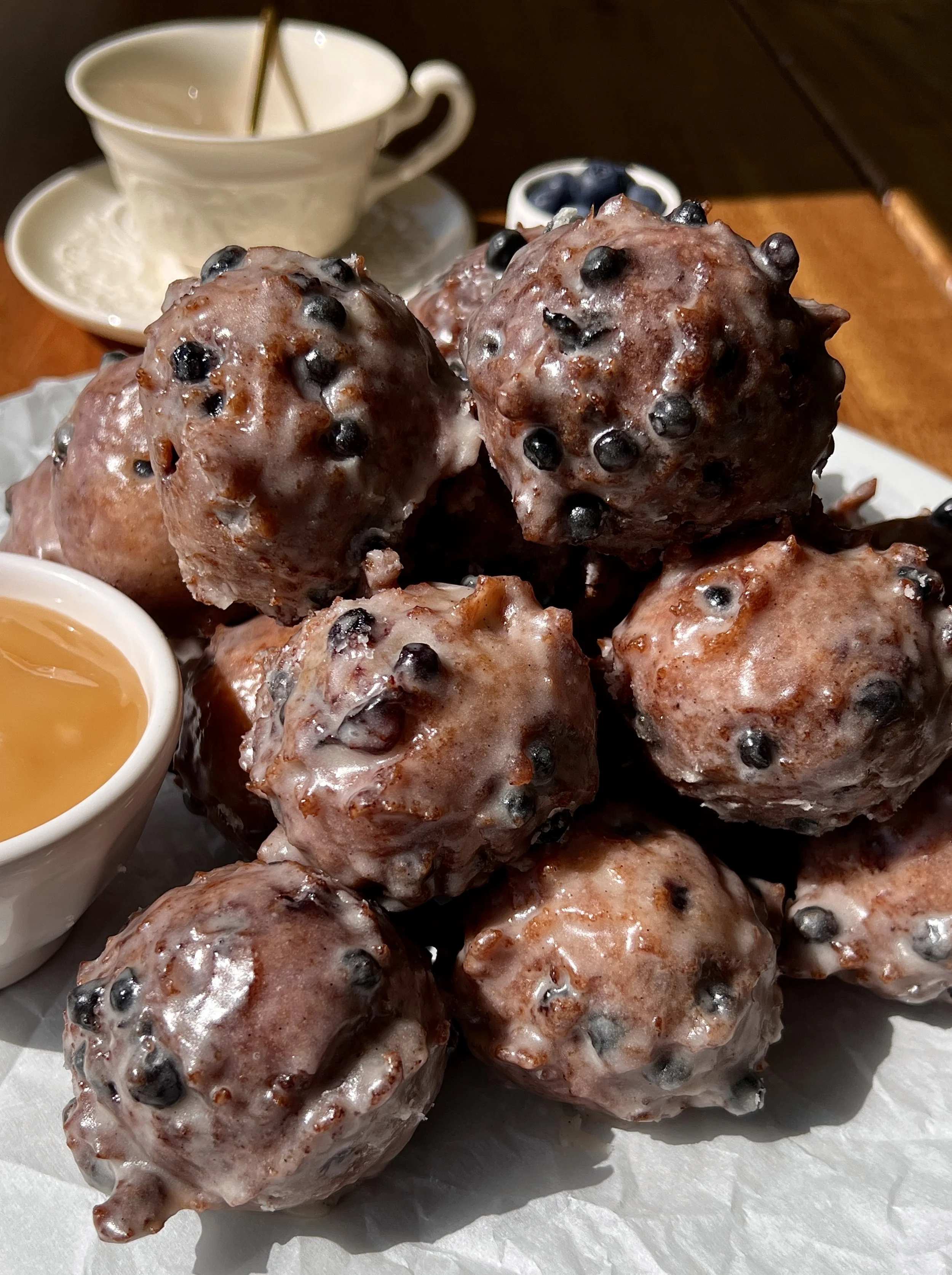 Pile of golden sourdough blueberry fritter bites coated in a silky vanilla bean glaze with bright juicy blueberries throughout served warm with a hot cup of coffee.