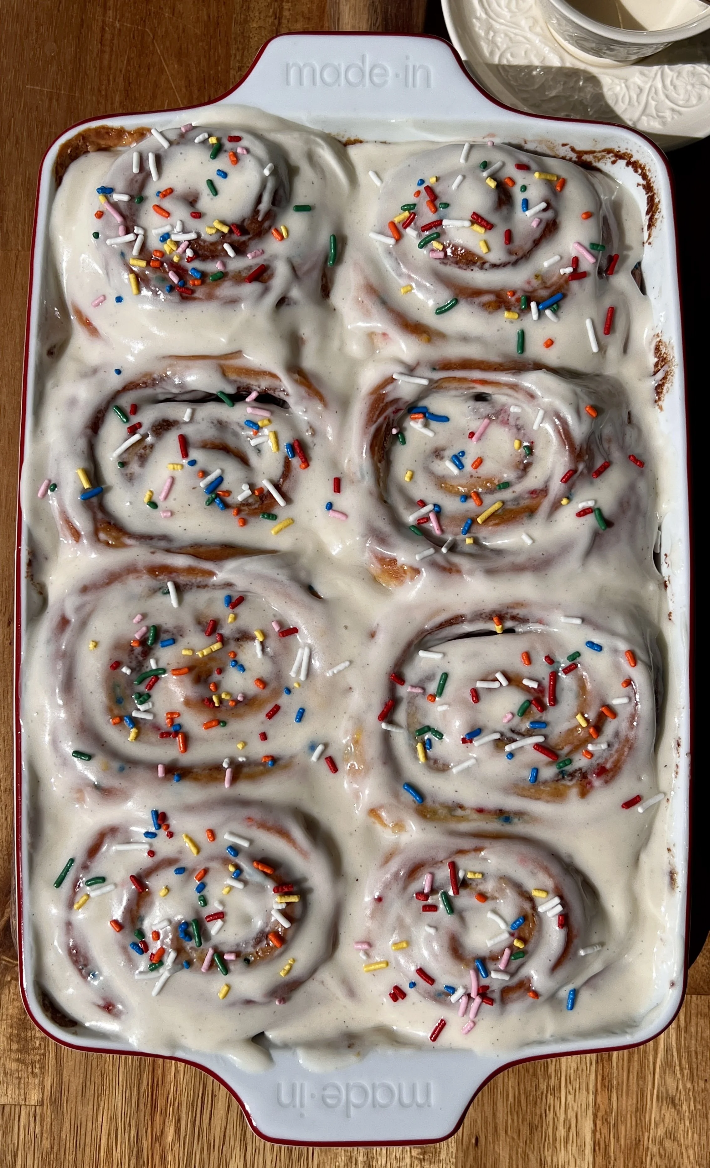 Pan of golden funfetti cinnamon rolls with sticky caramelized edges topped with smooth cream cheese frosting and extra rainbow sprinkles.