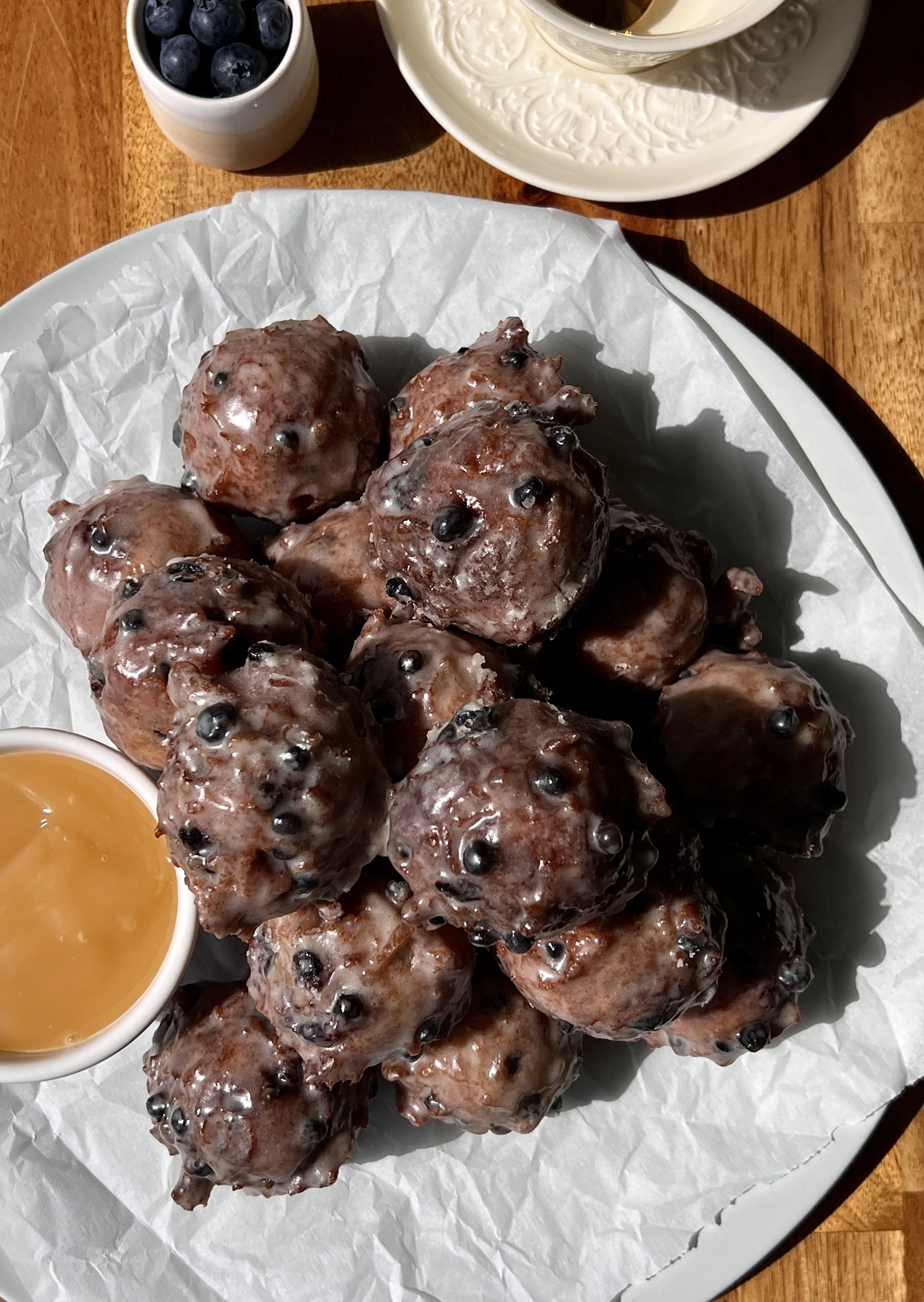 Pile of golden sourdough blueberry fritter bites coated in a silky vanilla bean glaze with bright juicy blueberries throughout served warm with a hot cup of coffee.