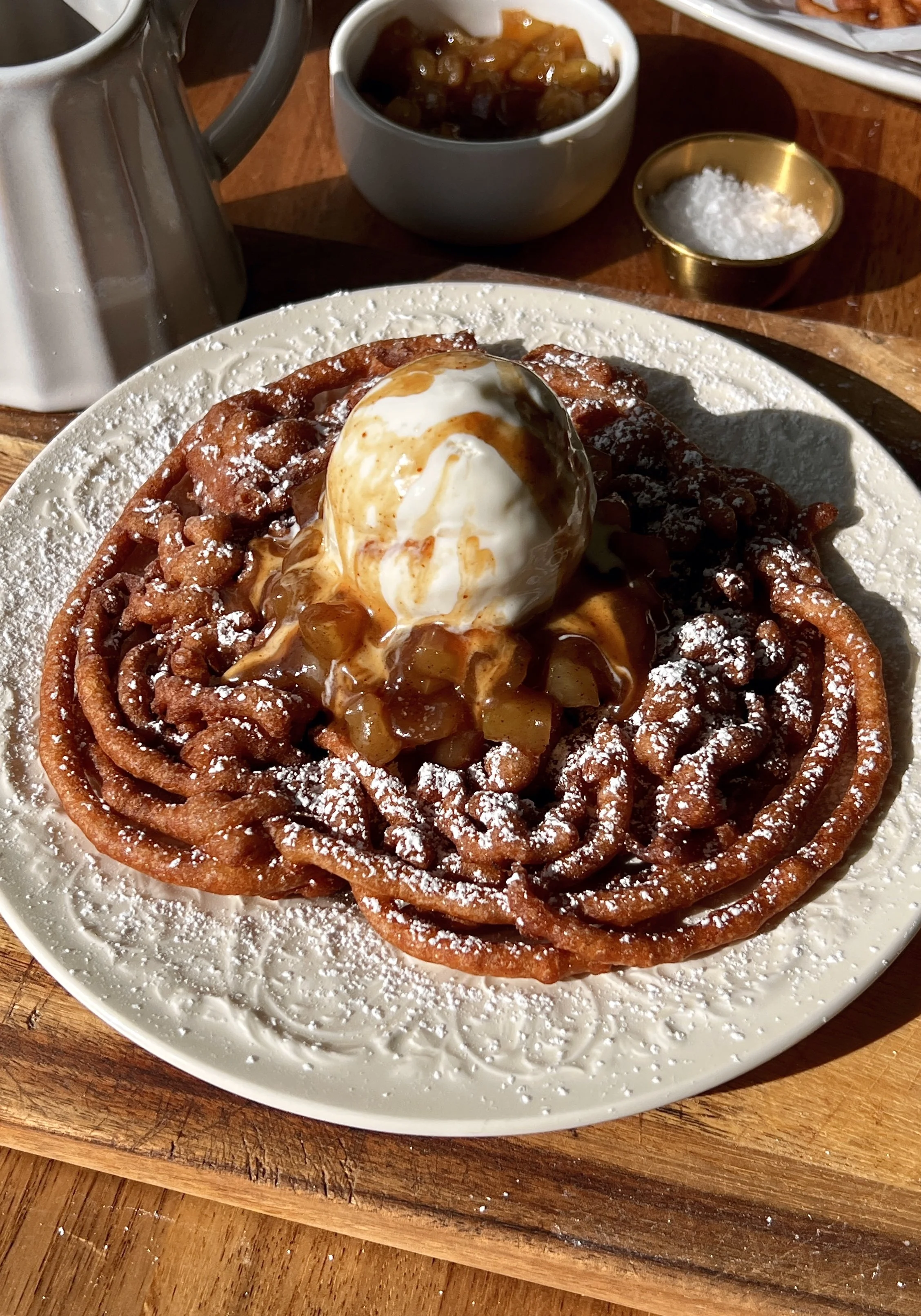 Apple cider funnel cakes perfectly golden and crisp served warm with a dusting of powdered sugar a gooey apple topping a scoop of vanilla ice cream and a drizzle of salted caramel.