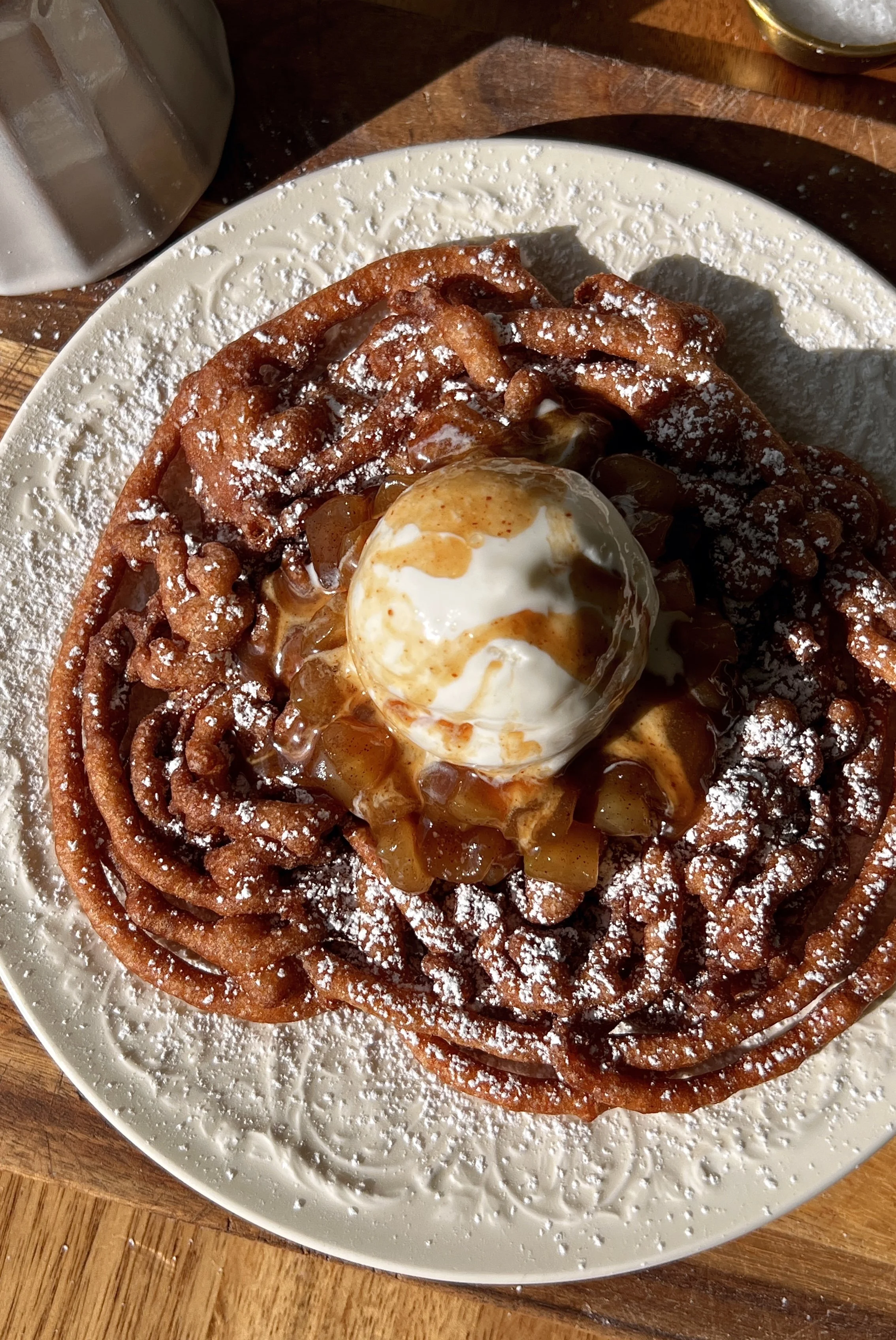 Apple cider funnel cake being topped with a gooey apple topping a scoop of vanilla ice cream and a drizzle of salted caramel served warm as the ultimate fall fair food at home.