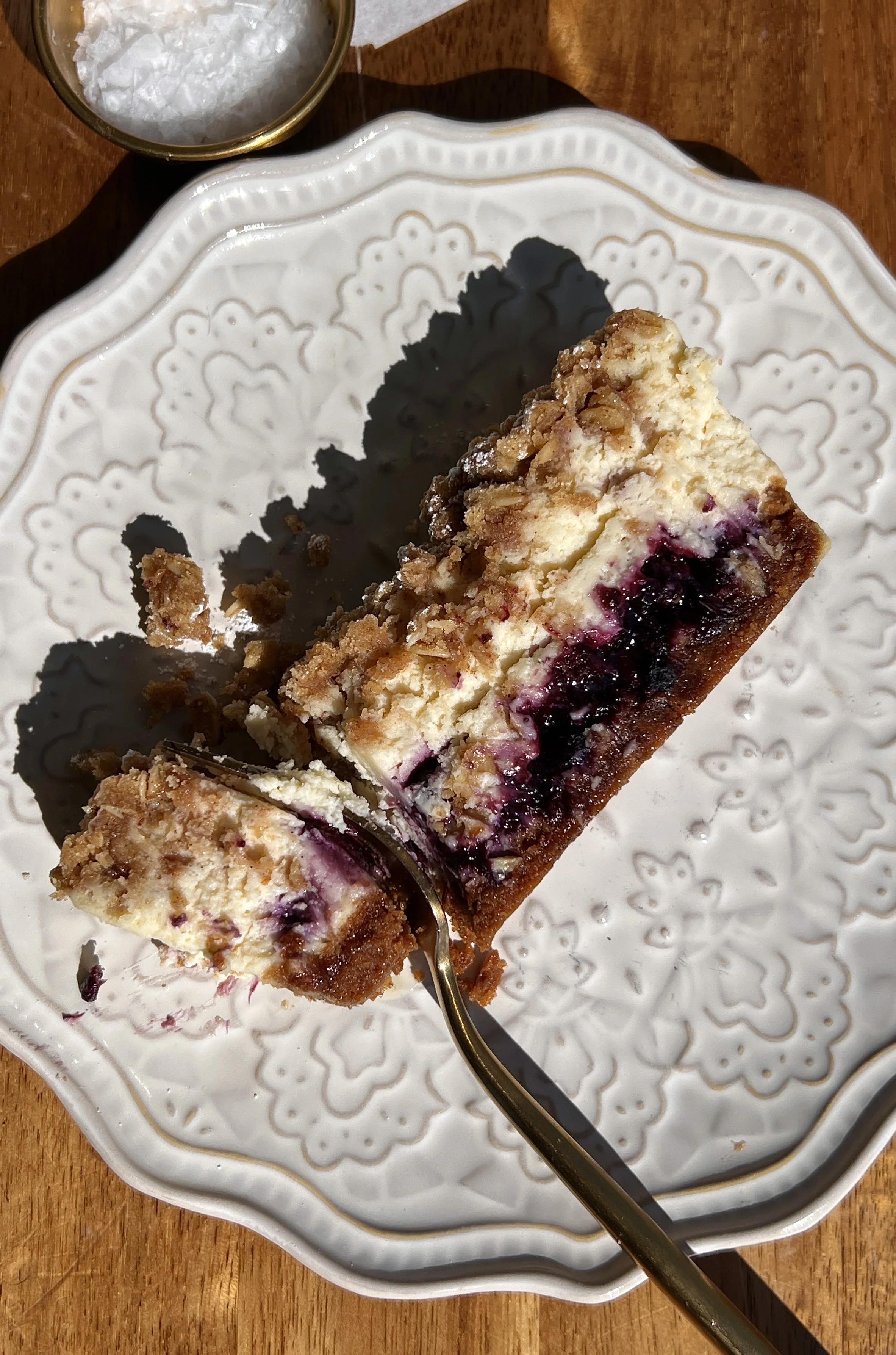 Blueberry crumble cheesecake bars with a buttery Biscoff cookie crust, tangy vanilla bean cheesecake filling, homemade blueberry compote, and golden crumble topping served on a plate.
