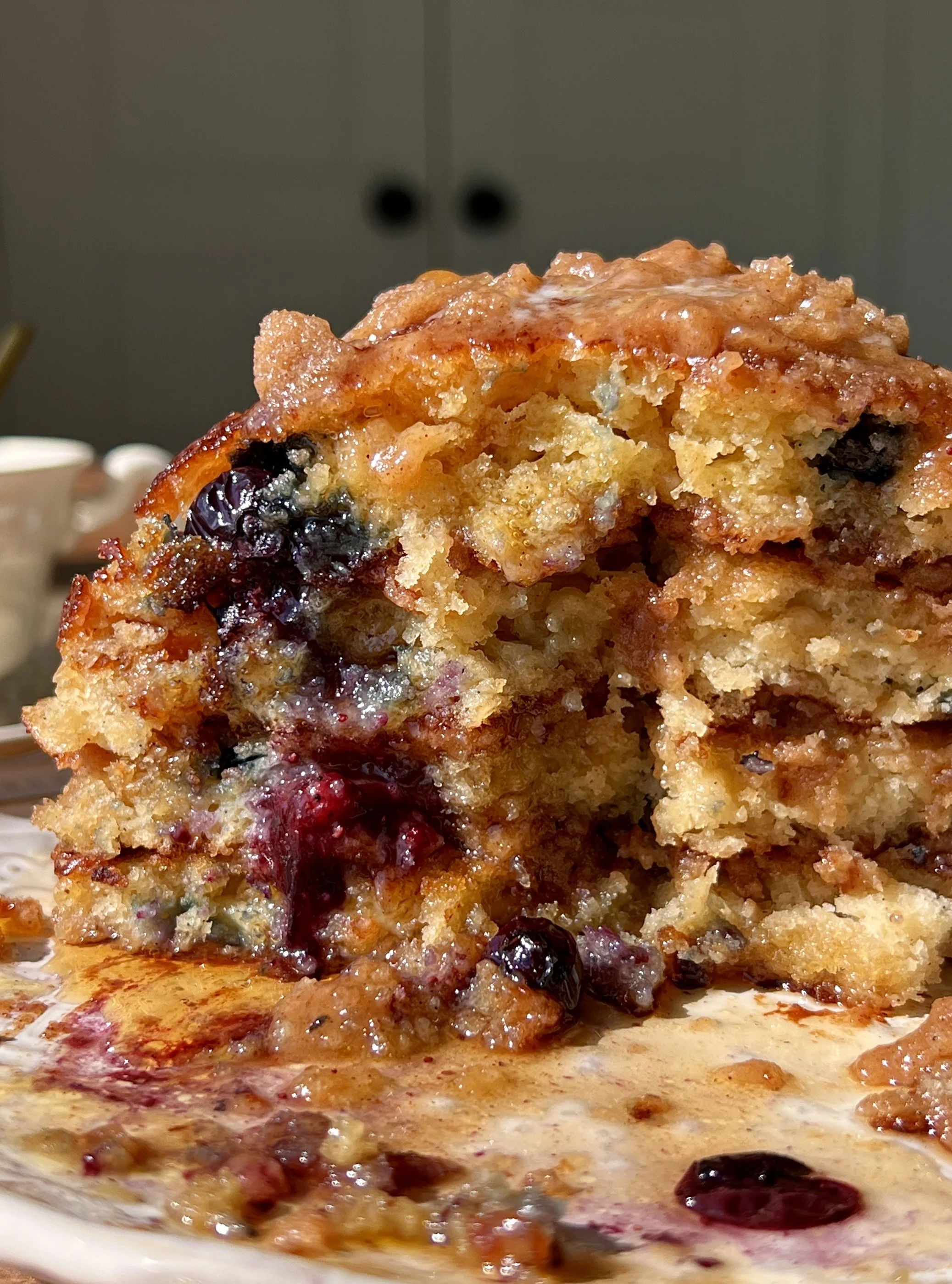 Stack of golden brown butter blueberry muffin pancakes with caramelized cinnamon streusel and juicy blueberries served warm with a pat of butter, maple syrup, and a hot cup of coffee.