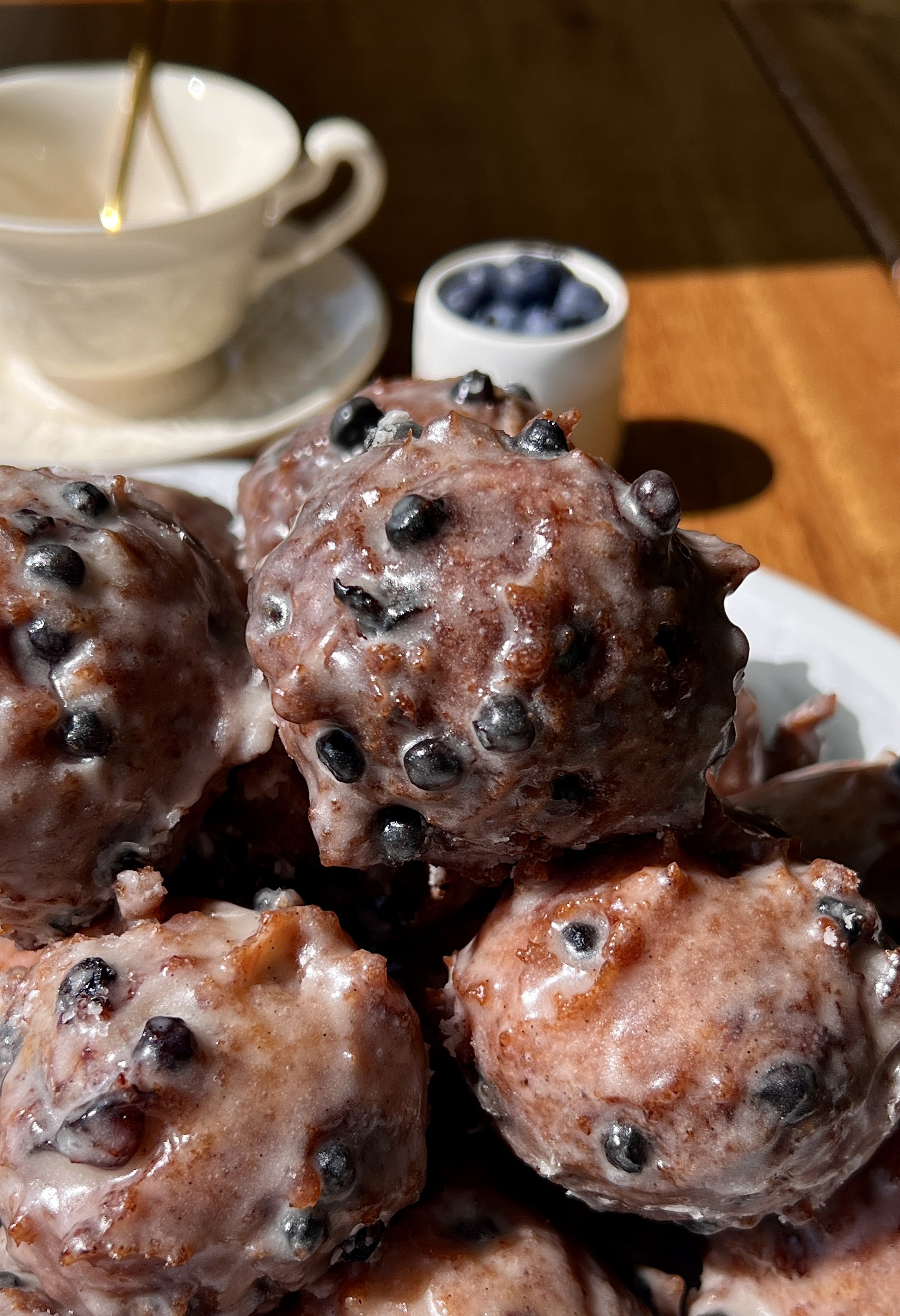 Pile of golden sourdough blueberry fritter bites coated in a silky vanilla bean glaze with bright juicy blueberries throughout served warm with a hot cup of coffee.