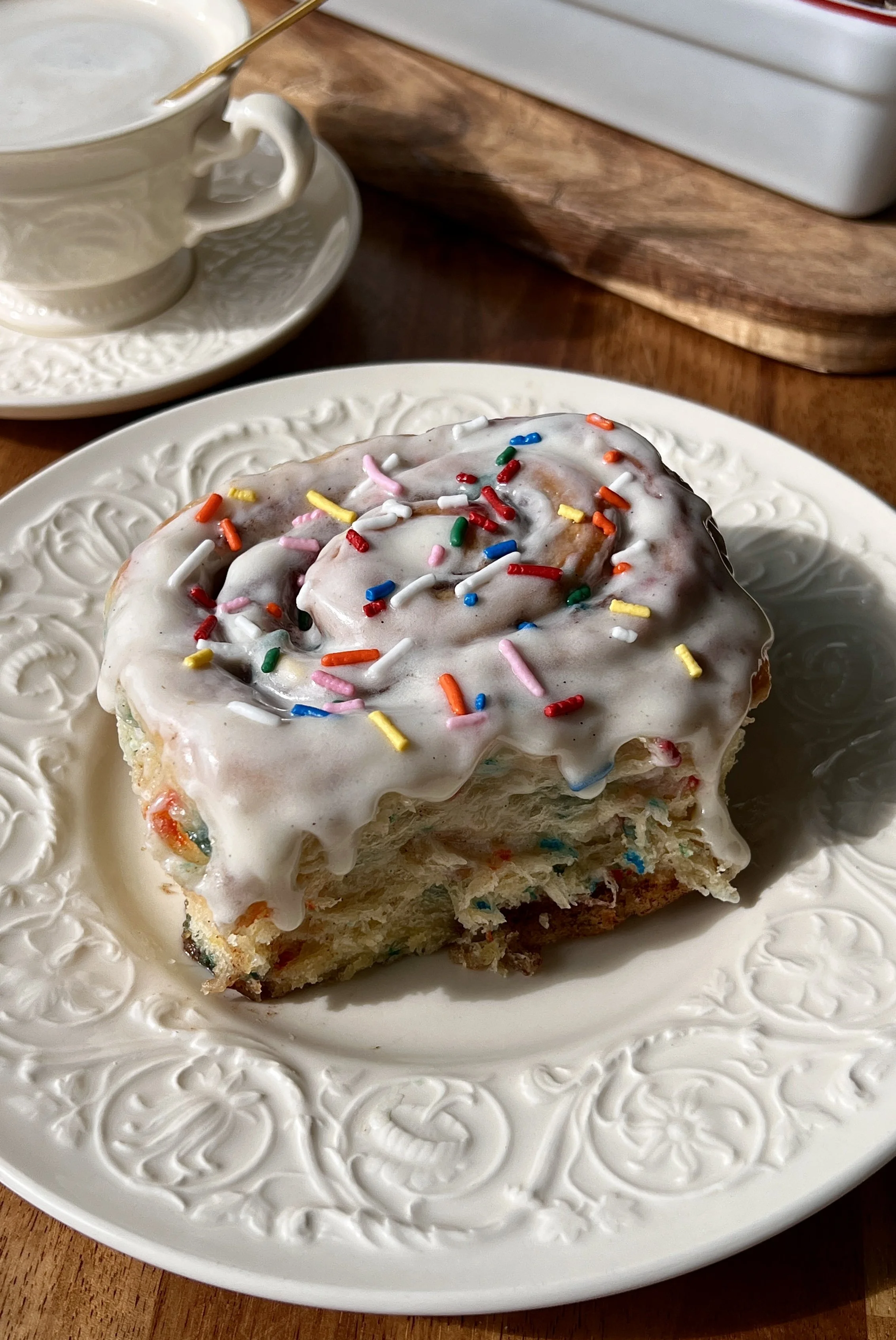 Funfetti cinnamon rolls with cream cheese frosting and rainbow sprinkles served warm with a hot cup of coffee.
