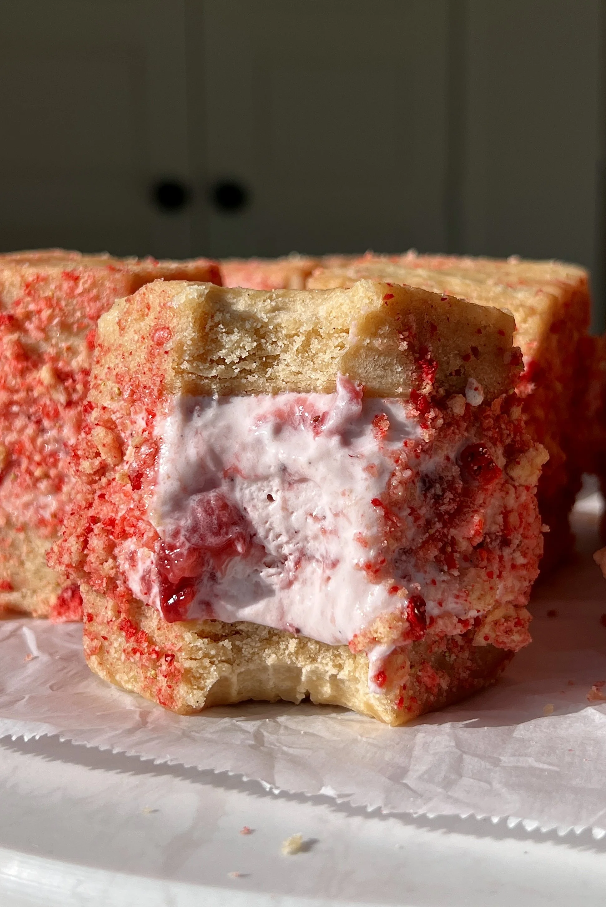 Strawberry shortcake shortbread ice cream sandwich with a bite taken out showing the soft buttery shortbread layers with rich and creamy homemade no-churn strawberry ice cream and strawberry shortcake crunch coating inside.