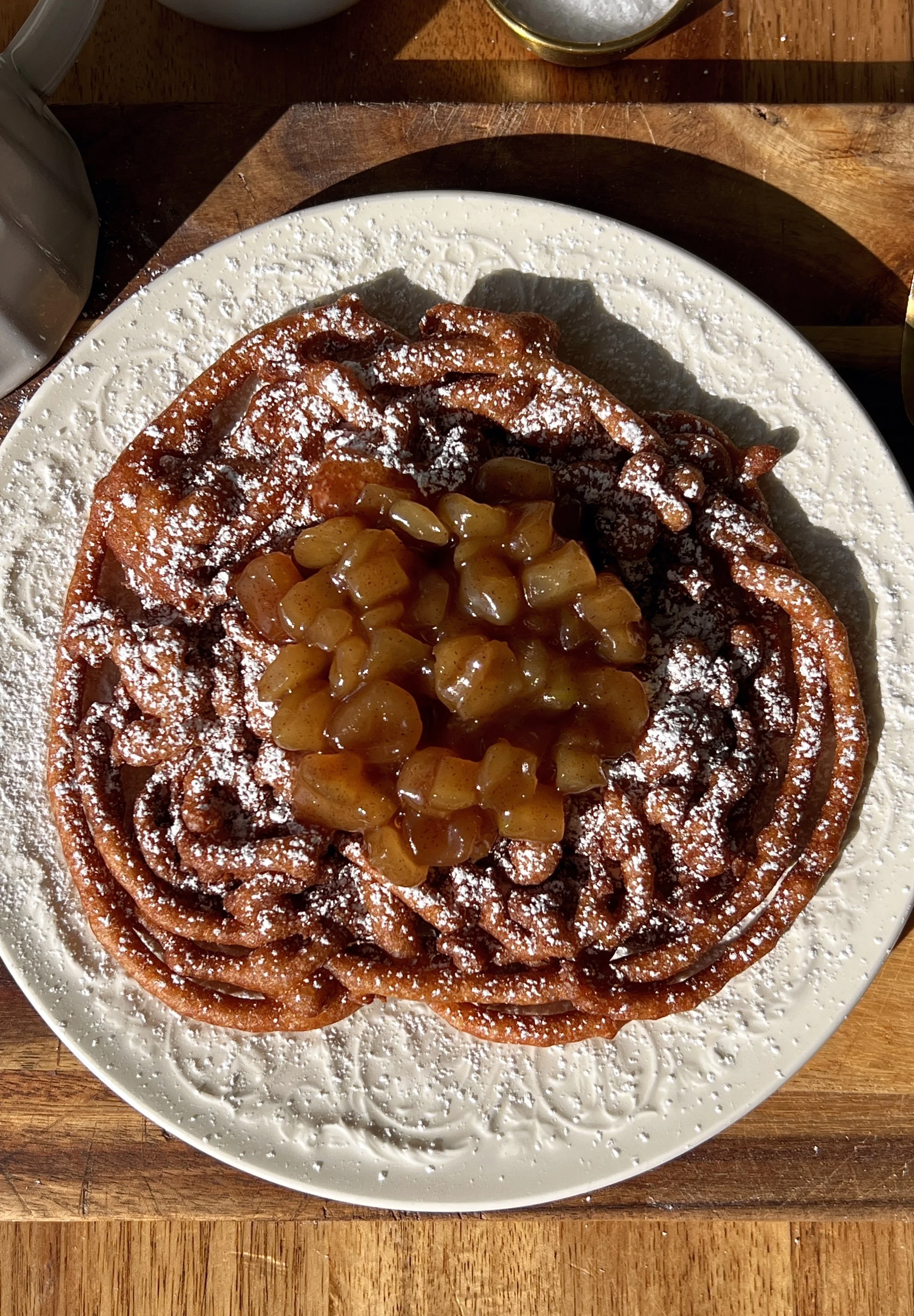 Apple cider funnel cake being topped with a gooey apple topping a scoop of vanilla ice cream and a drizzle of salted caramel served warm as the ultimate fall fair food at home.