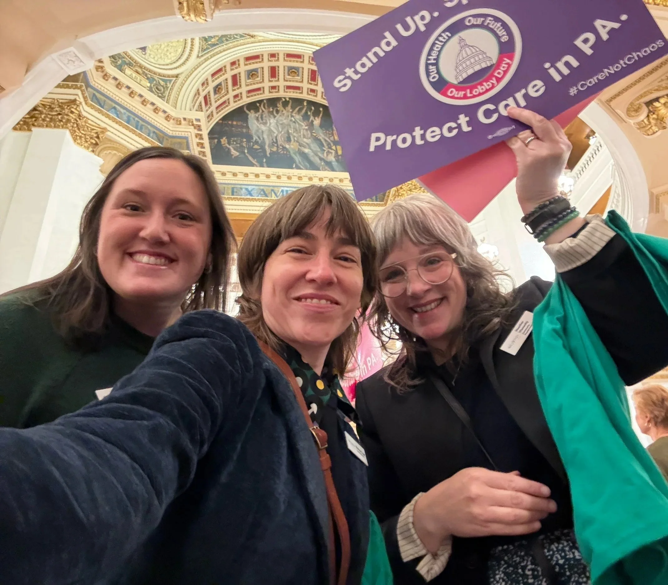 Three women smiling at the camera. One is holding a sign that says "Protect Care in PA"