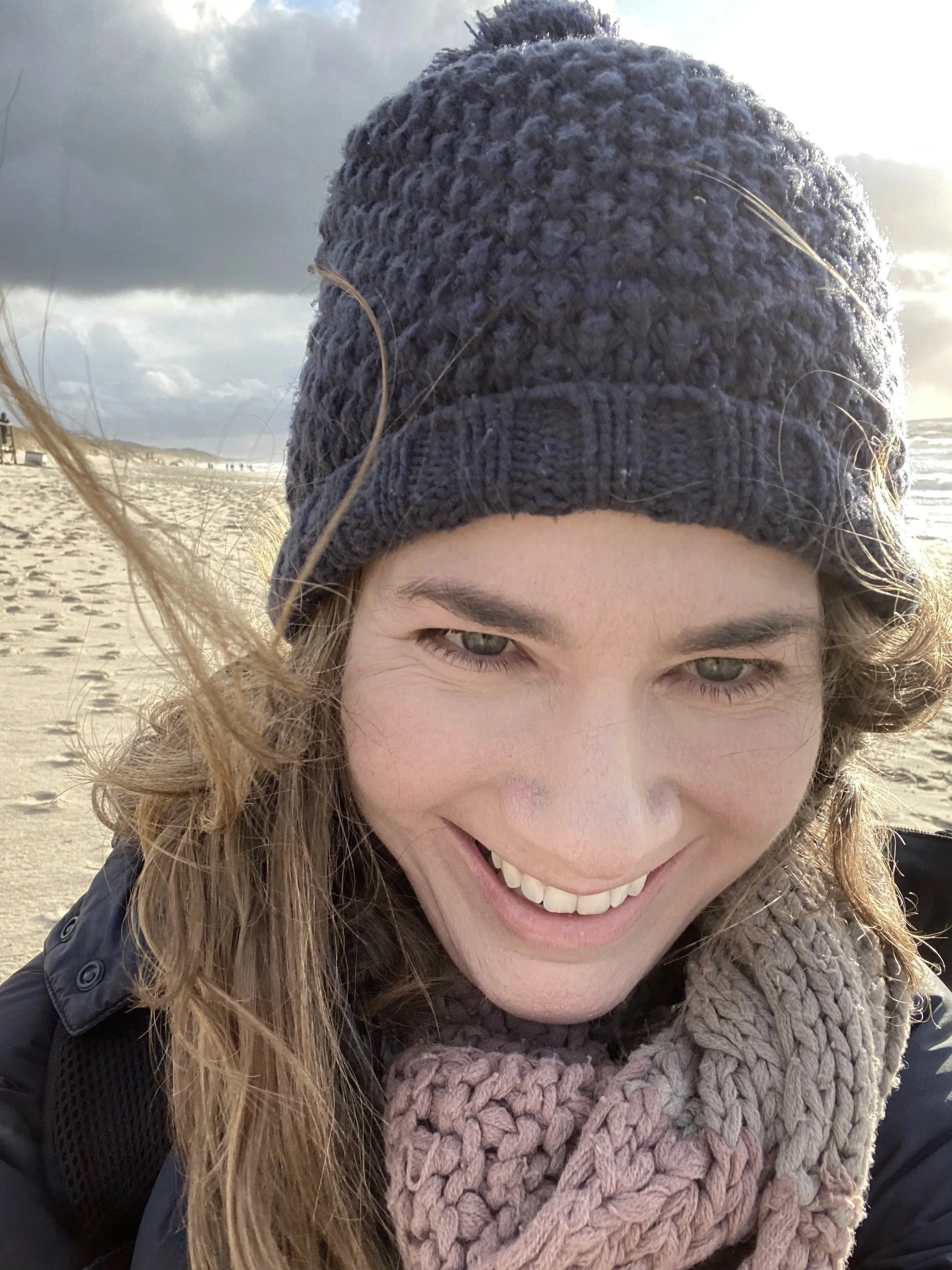 Lächelnde junge Frau mit langen, lockigen Haaren trägt eine graue Mütze, einen dicken Beanie, und einen dick gestrickten Schal. Im Hintergrund ein Strand bei bewölktem Himmel.