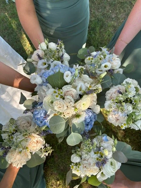 A top-down view of multiple white and light blue floral bouquets with green eucalyptus leaves, held by several people in green dresses on a grassy field during daytime.