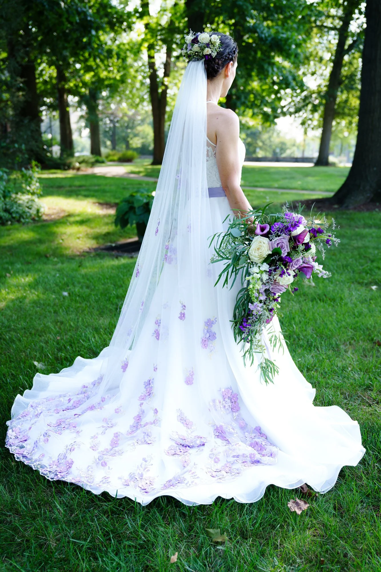 A bride in a white wedding dress with purple floral accents, holding a large purple and white bouquet, standing outdoors on grass with trees in the background.