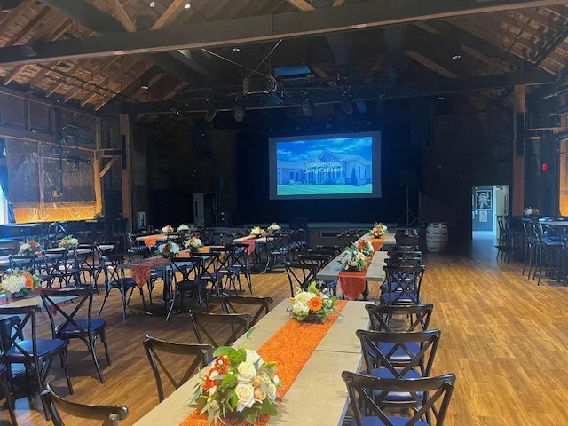 Event space with wooden floors, decorated tables with floral centerpieces, and a large screen at the front showing a city skyline.