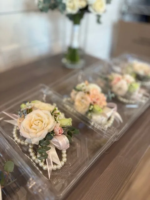 Wedding corsage with white roses, pink flowers, greenery, a pearl wrist band, and ribbon, displayed in a protective case on a wooden table