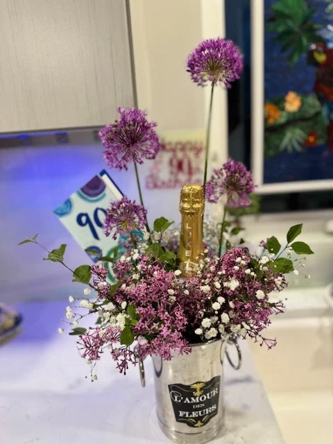 Small bouquet of pink and white flowers with greenery in a silver bucket with a champagne bottle and purple allium flowers