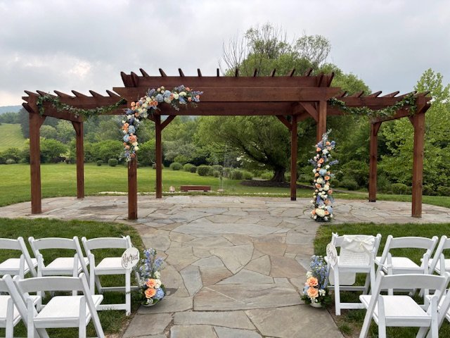 Outdoor wedding setup at Wind in the Willows Grantville, PA with a wooden pergola decorated with floral arrangements, white chairs, and a stone pathway in a lush green park.