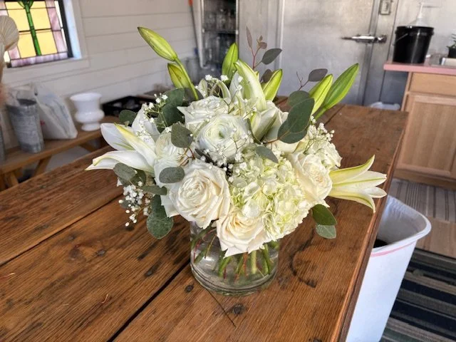 A bouquet of white roses, lilies, and other white flowers with green foliage in a glass vase on a wooden table.