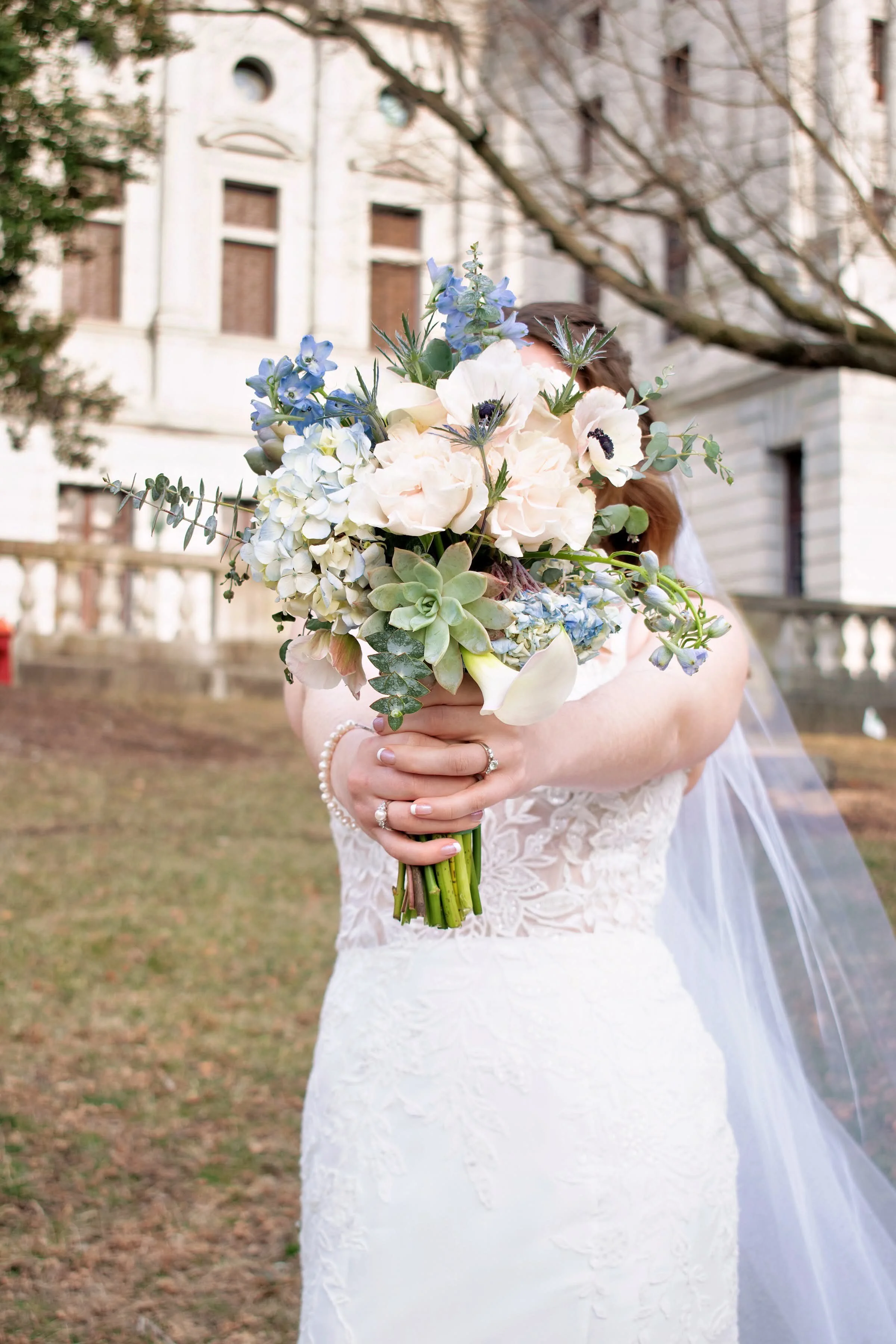 A bride holding a large bouquet of cream, white, blue flowers, and greenery in front of her face outdoors.