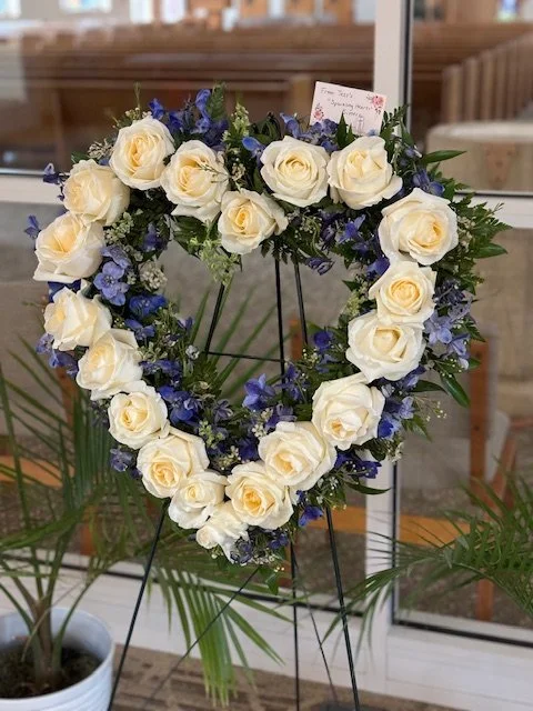 Heart-shaped floral wreath with white roses and purple flowers on a black metal stand, placed indoors near a window.