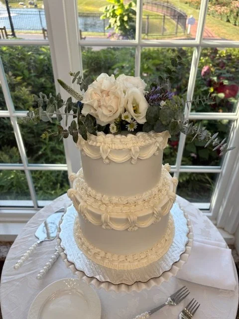 Three-tier white wedding cake with floral decoration of white rose, ranunculus, and eucalyptus, placed on a table near a window overlooking a garden.
