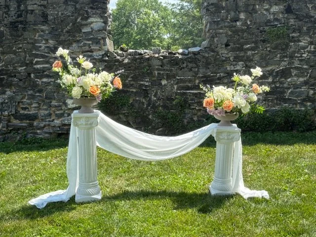 Wedding arch with two white columns adorned with flowers and draped white fabric, set outdoors on green grass near a stone wall.