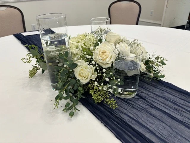 A centerpiece with white roses, baby's breath, and greenery on a navy table runner with three votives for floating candles on a white table.