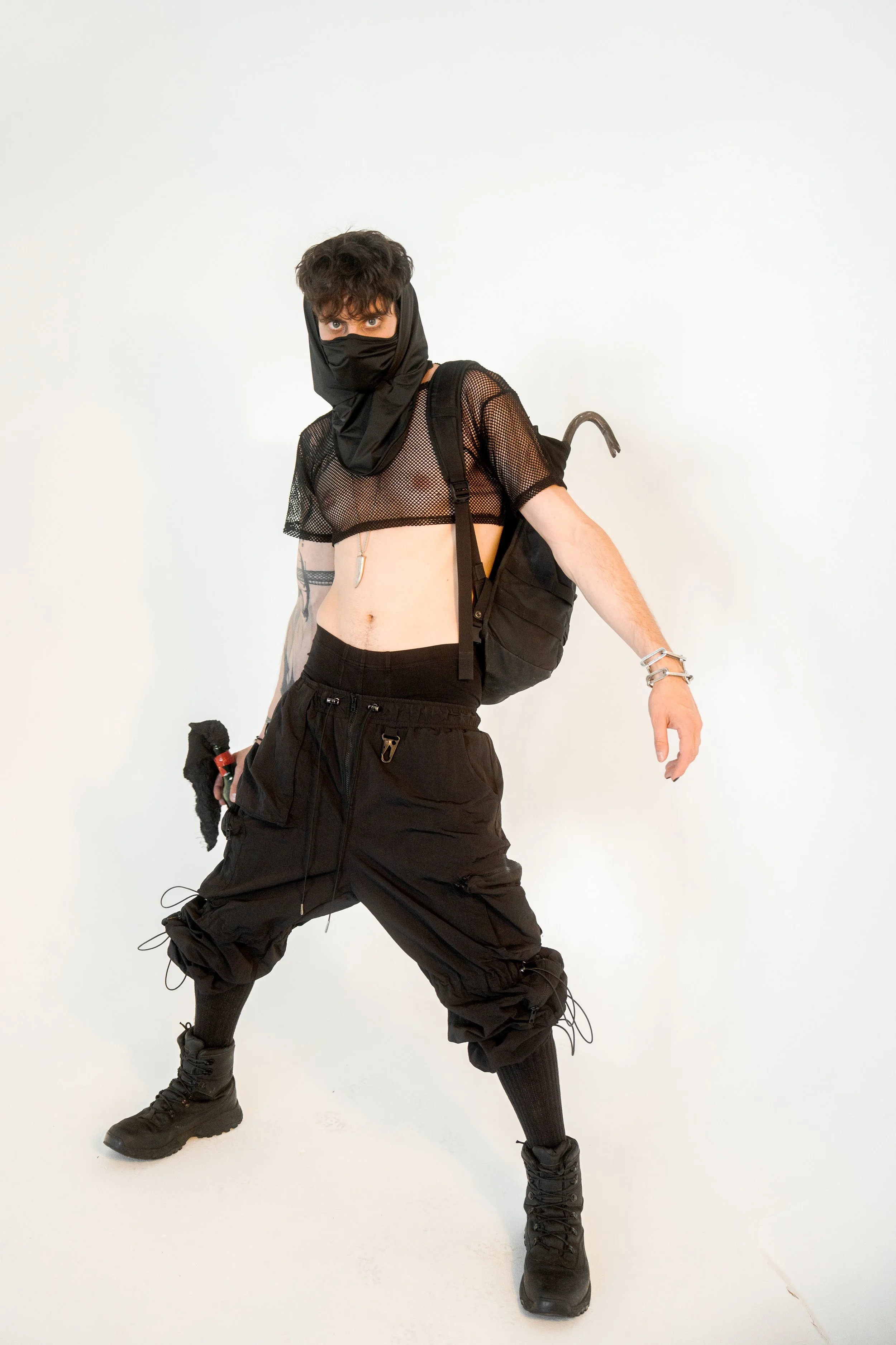 XINIX dressed in black clothing with face mask, standing against a white background, holding a small object in one hand, with a backpack over one shoulder.
