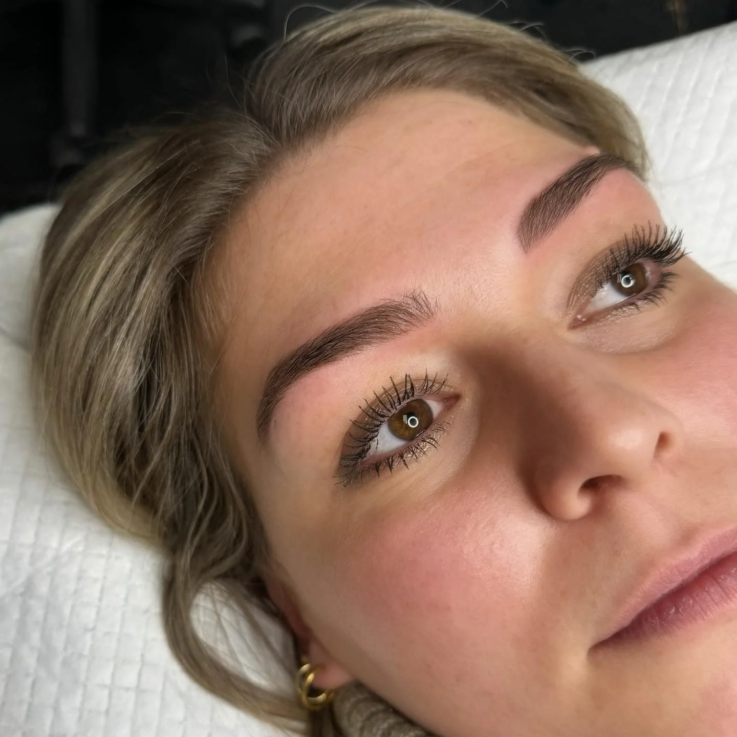 Une femme avec des sourcils bien définis, des cils longs et maquillage subtle, allongée sur un lit avec une serviette blanche derrière elle.
