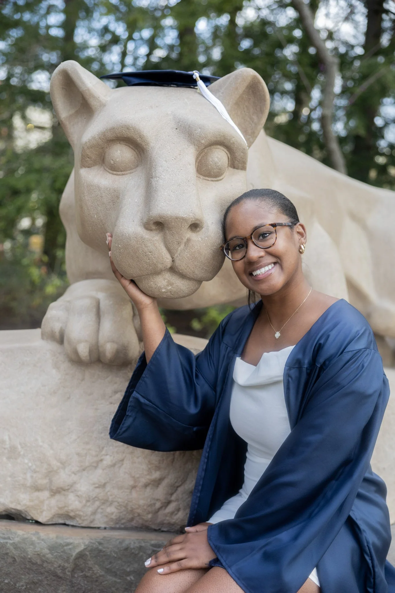 A woman in graduation attire, wearing glasses and a jewelry, sitting outdoors with a large lion statue wearing a graduation cap. She is smiling and holding the lion's face gently.