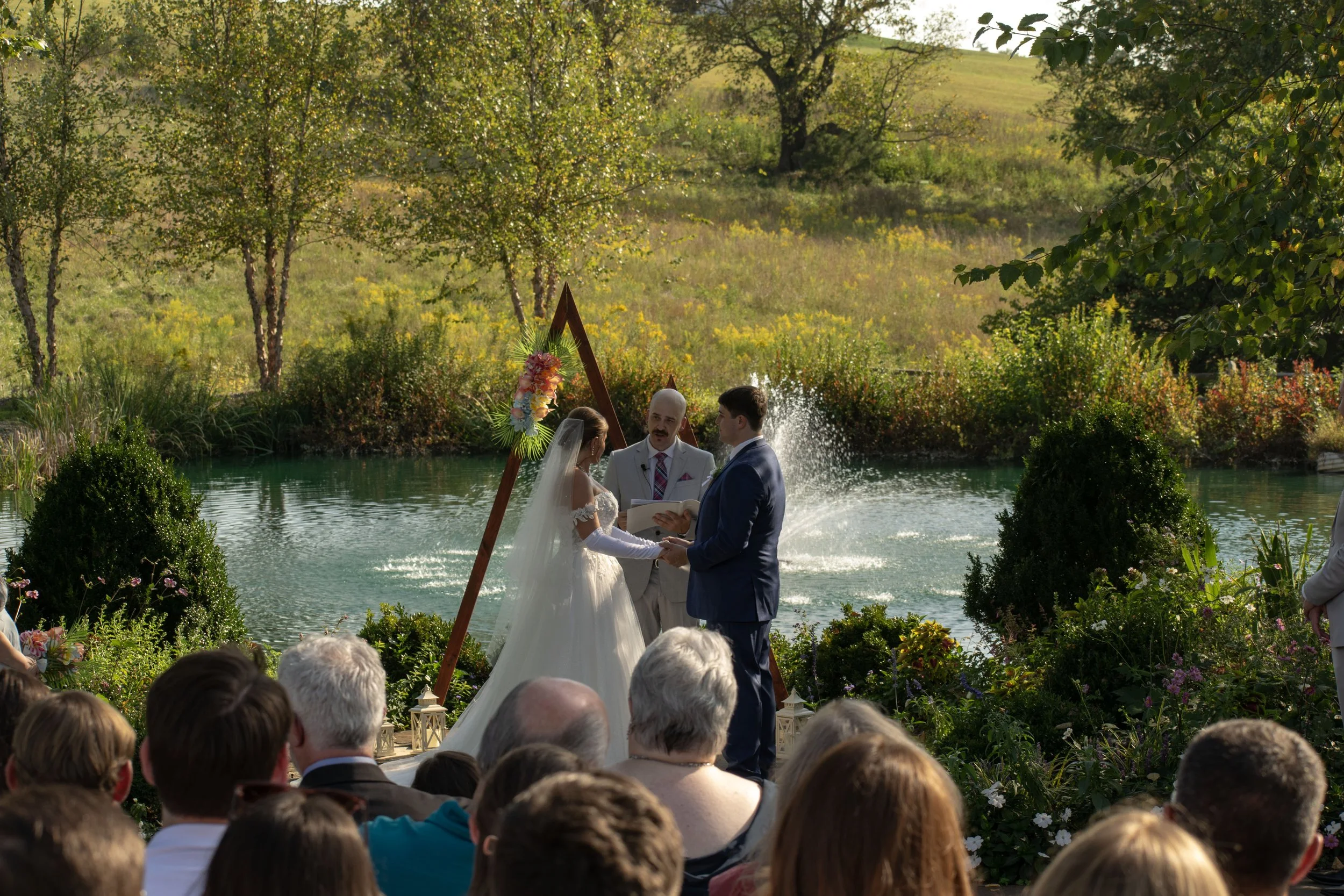 A wedding ceremony outdoors by a pond with trees and hills in the background, featuring a bride and groom exchanging vows in front of officiant, surrounded by seated guests.