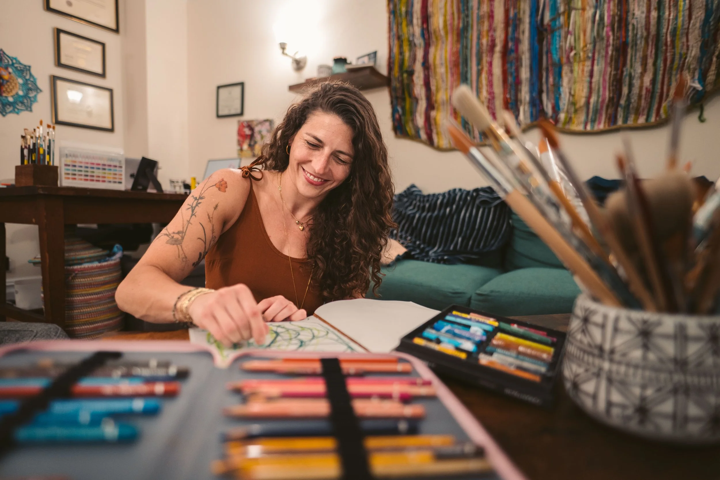 Abby Bramnick, Manhattan Art Therapist, with curly hair smiling and drawing with colored pencils at a table in a cozy living room, surrounded by art supplies and colorful decorations.