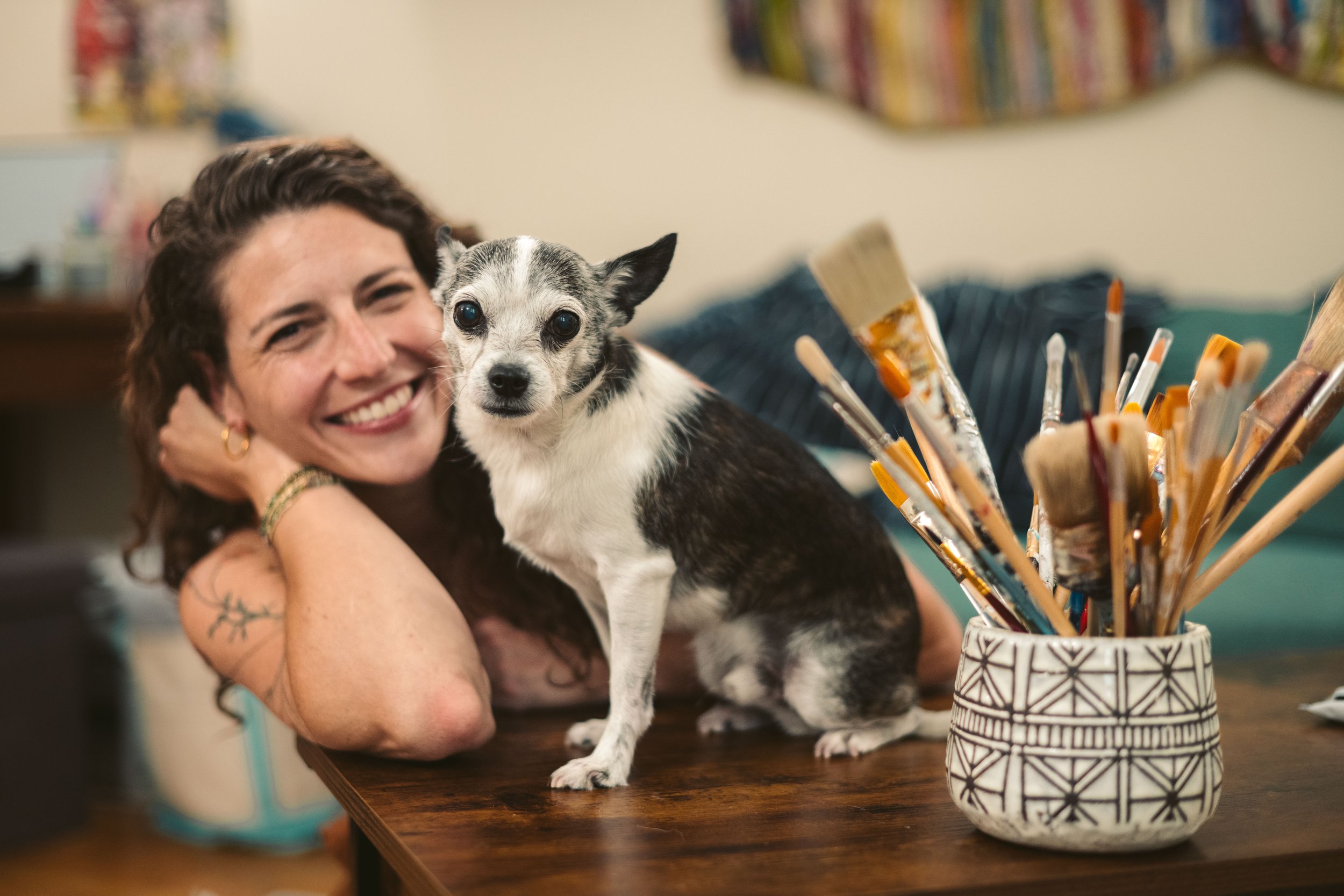 Abby Bramnick, Manhattan Art Therapist and Eating Disorder Therapist, smiling and lying on a wooden table with a small black and white dog with pointy ears, in an art studio with paintbrushes in a decorative container nearby.